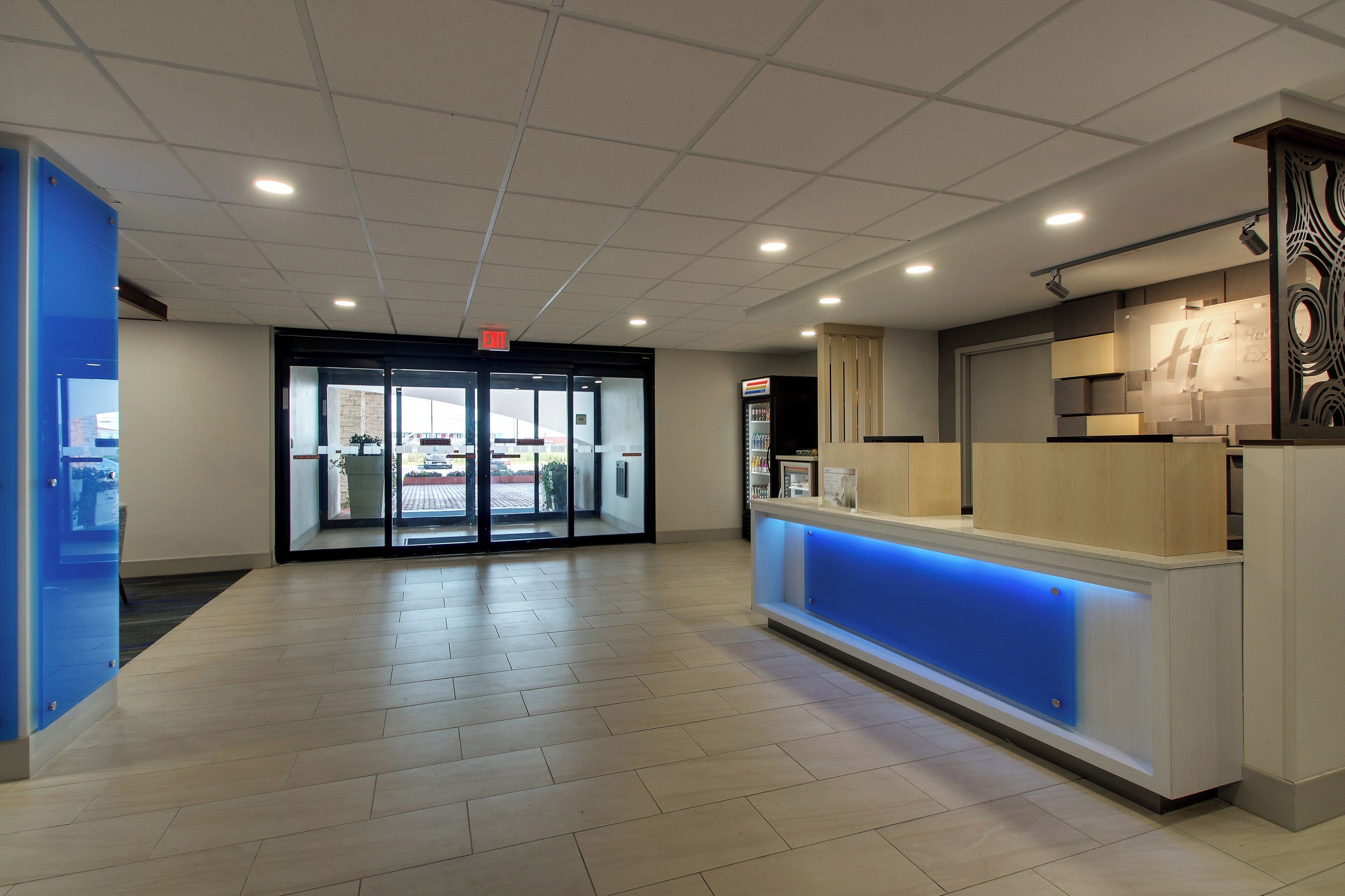 Zona de recepción de hotel con puerta de cristal, mostrador de recepción con iluminación azul y máquina expendedora.