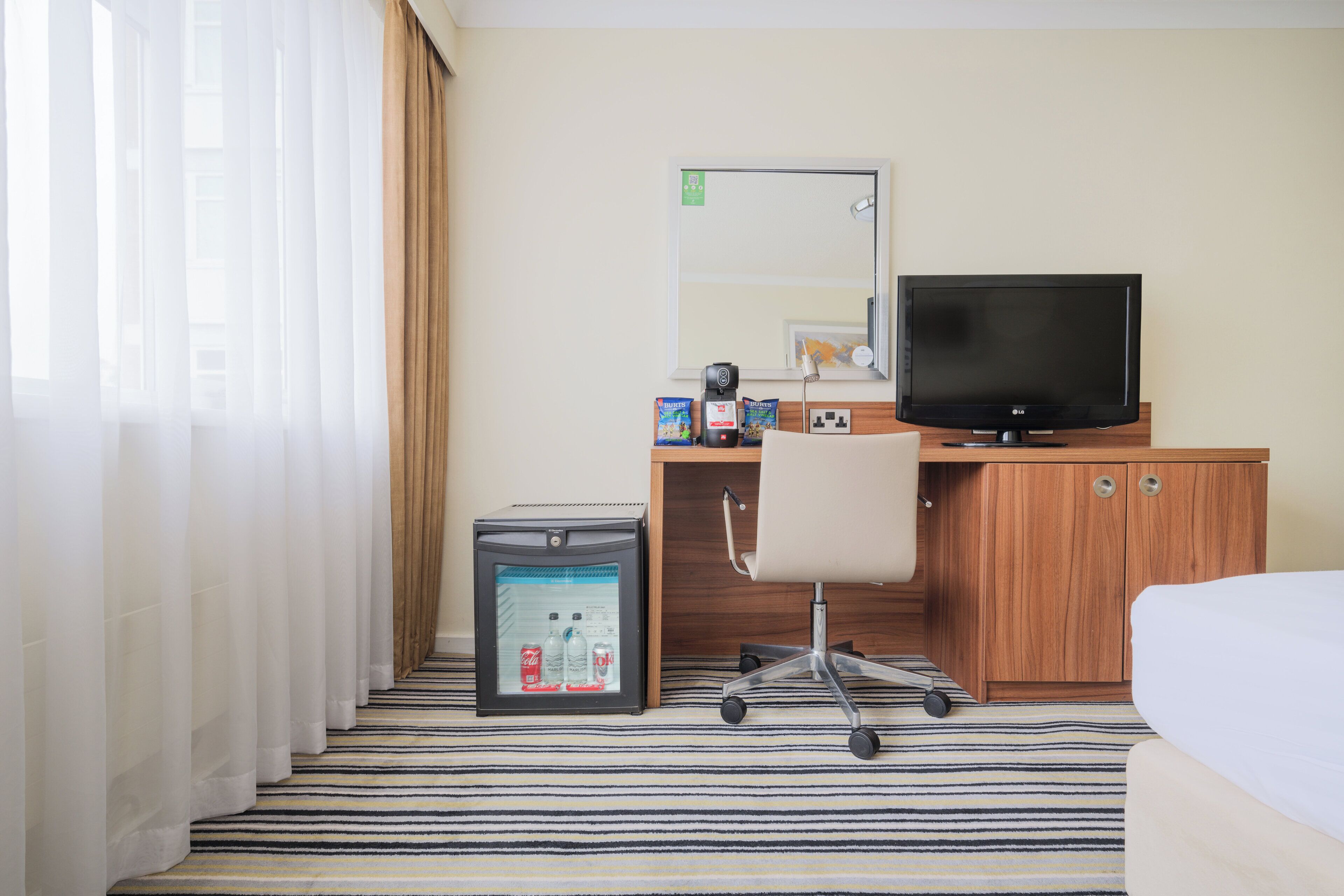 A hotel room with a bed, a desk with a computer and a television, a small refrigerator, and a window with curtains.
