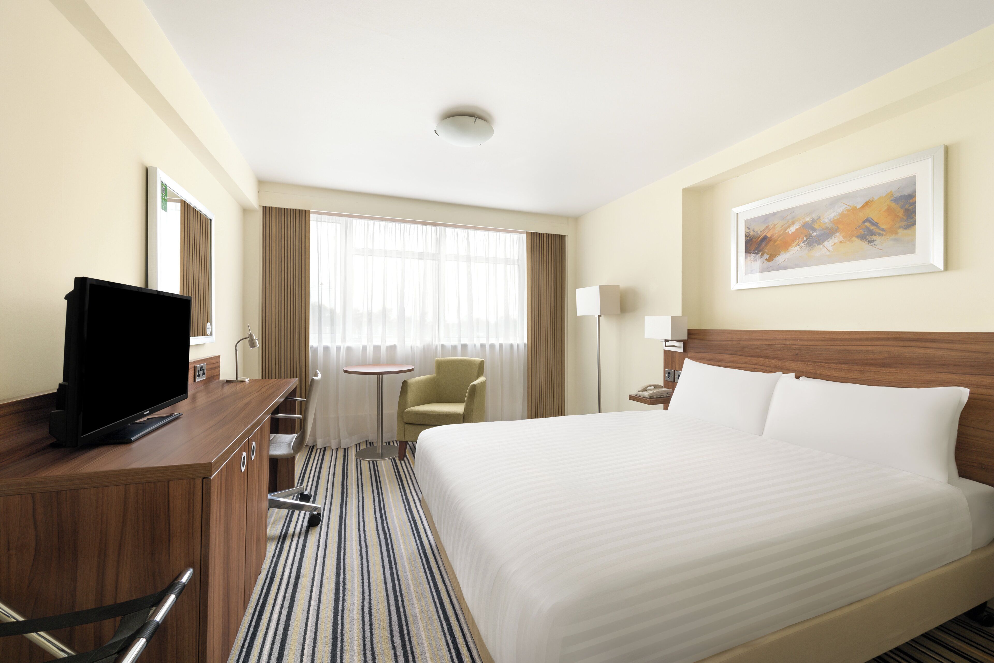 Habitación de hotel con cama, escritorio con televisor, una silla, una mesita y una ventana con cortinas.