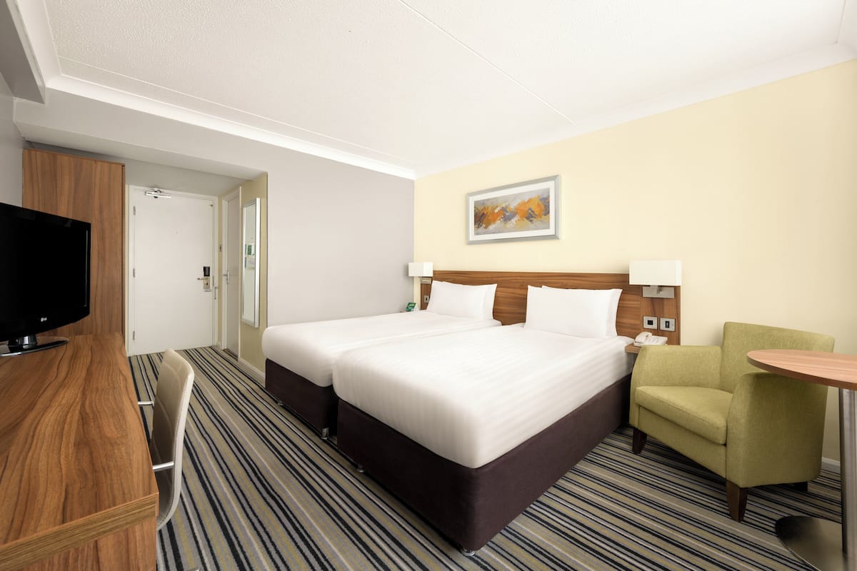 Ein Hotelzimmer mit zwei Betten, einem Fernseher, einem Schreibtisch, einem Sessel und einem kleinen Tisch.
