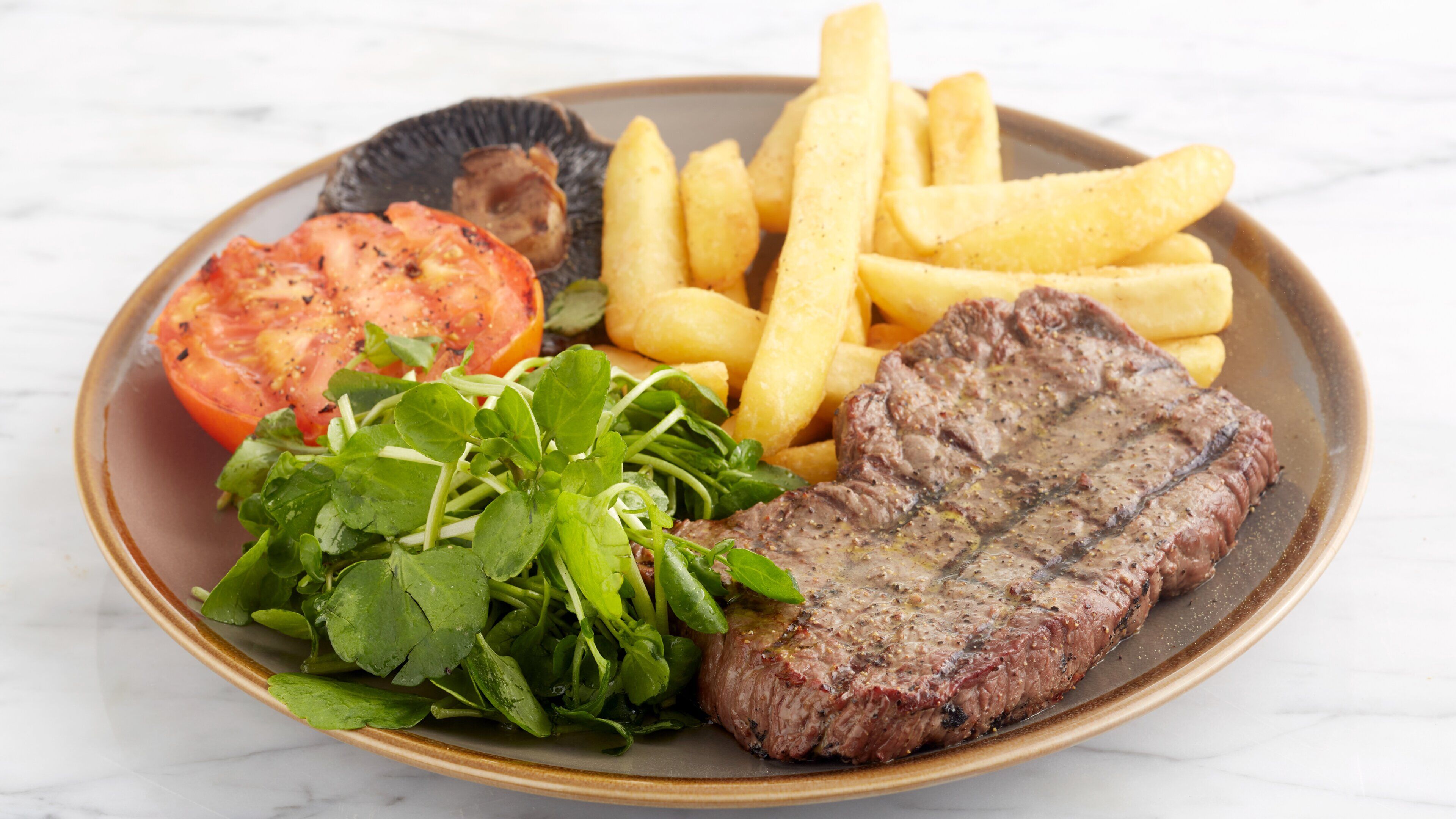 Un plato con un bistec, papas fritas, una rodaja de tomate y vegetales verdes.