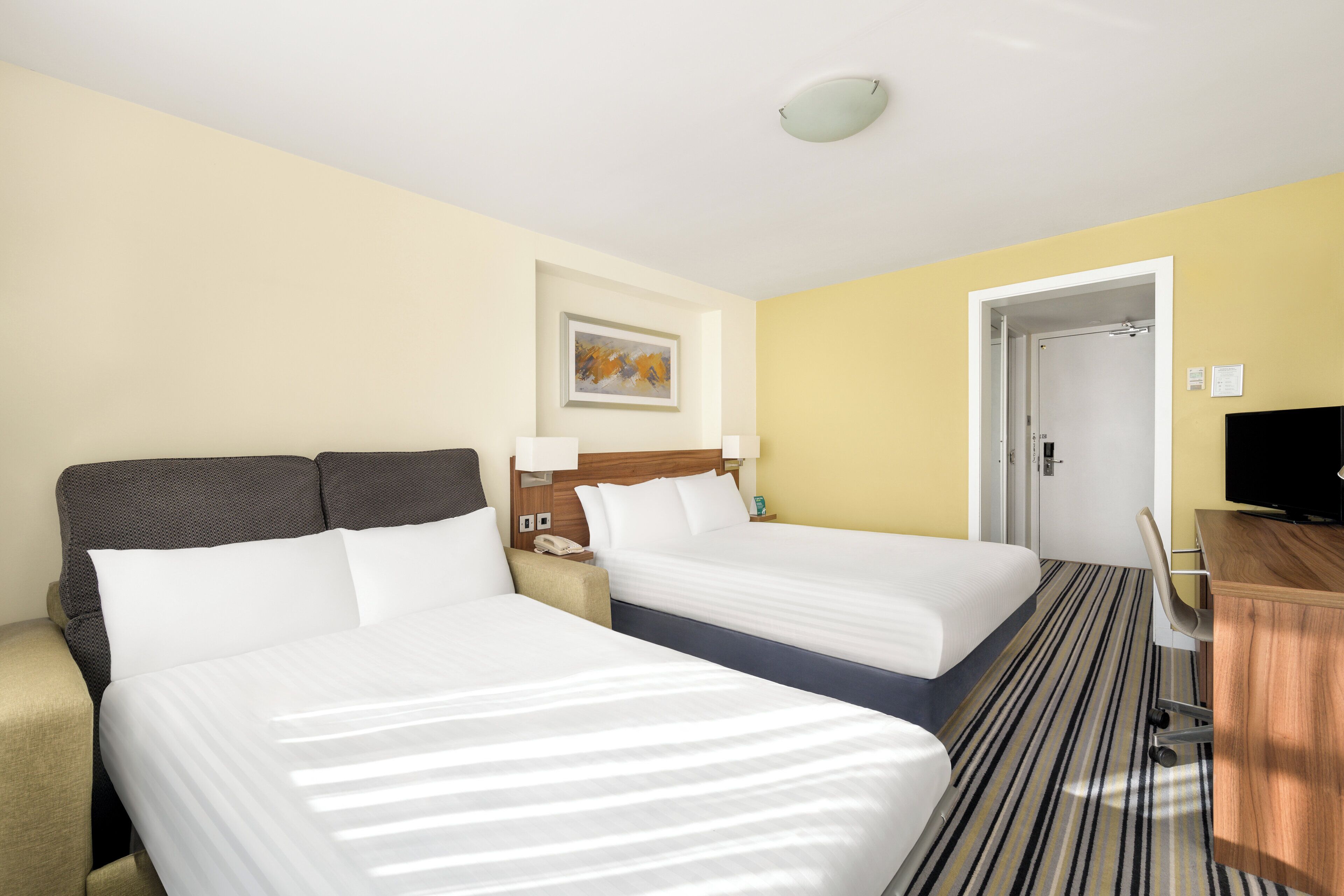 A hotel room with two beds, a sofa, a desk, a television, and a painting on the wall.