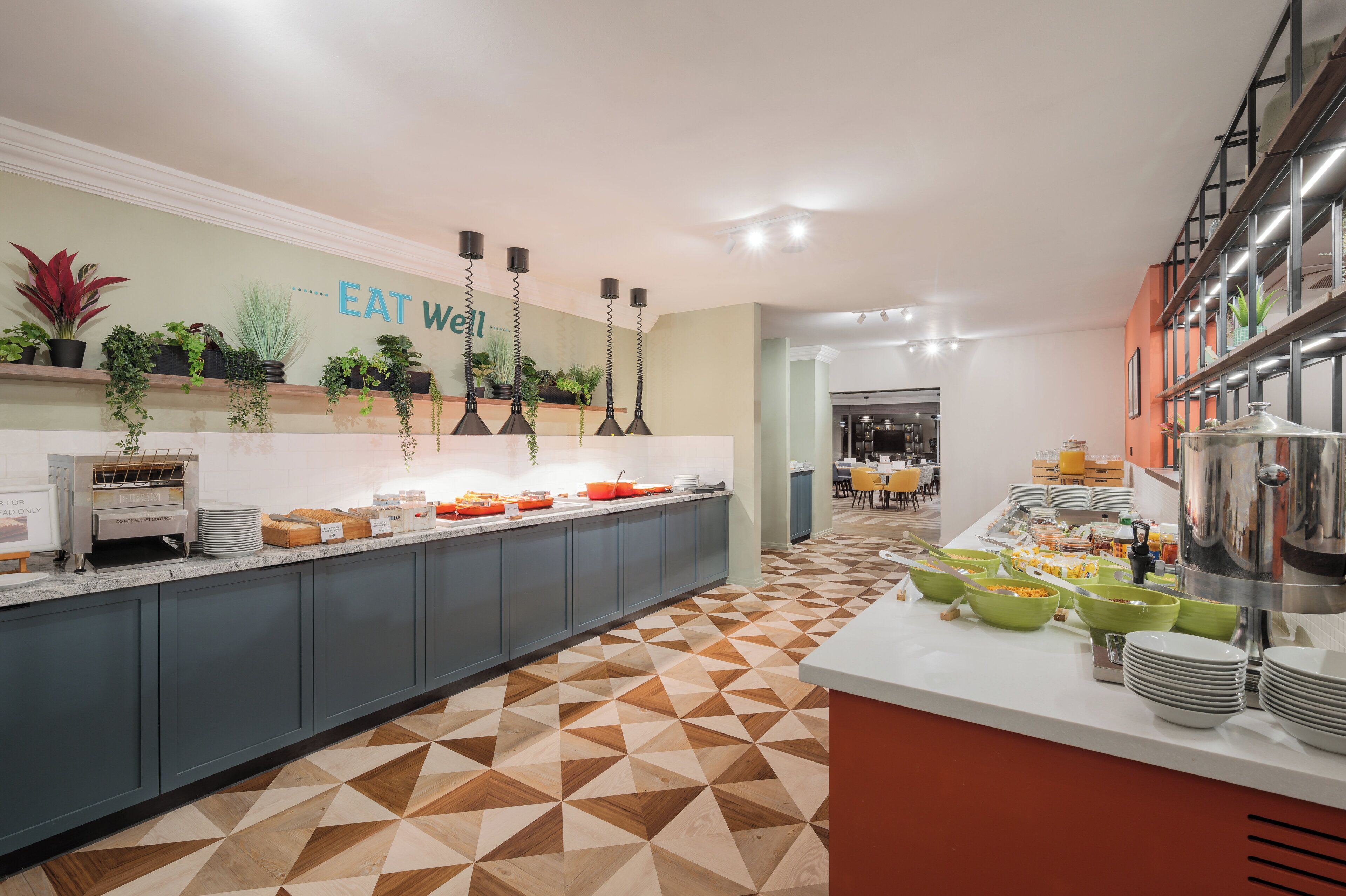 A modern kitchen with a geometric patterned floor, a buffet setup, and a sign that reads "EAT WELL".
