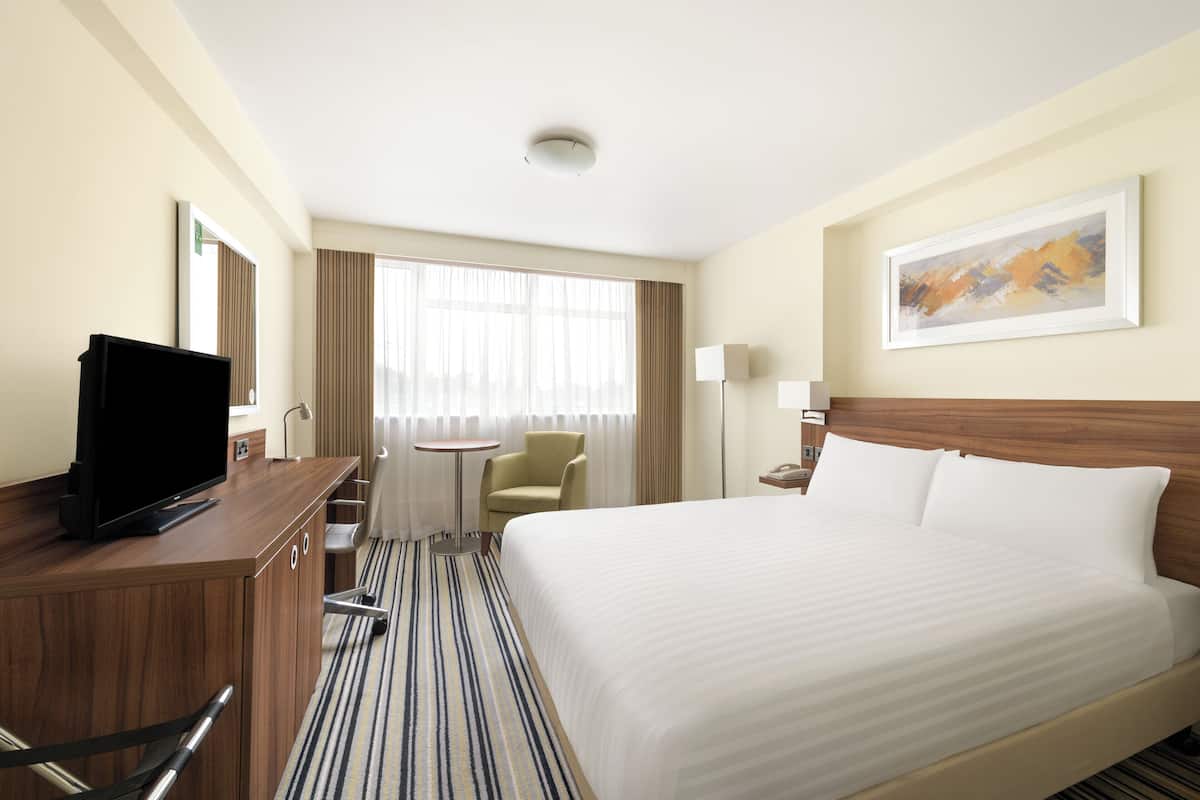Ein Hotelzimmer mit einem Bett, einem Schreibtisch mit Fernseher, einem Sessel, einem kleinen Tisch und einem Fenster mit Vorhängen.