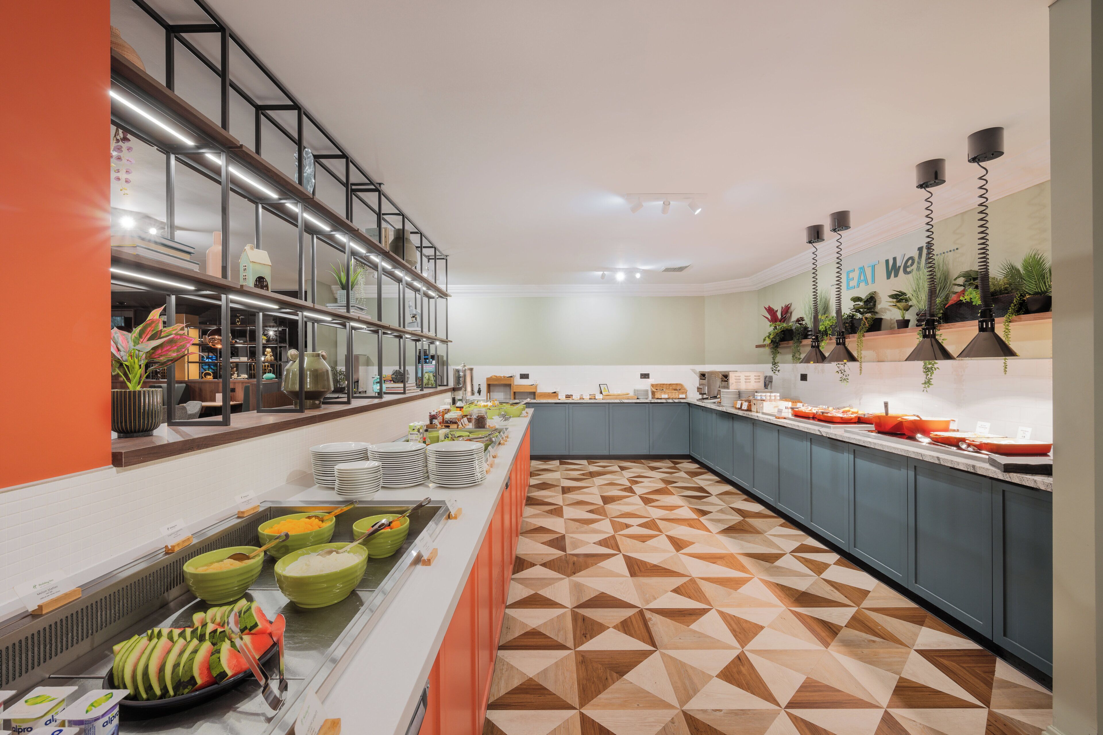 A modern buffet with a geometric patterned floor, a variety of food items, and a 'EAT Well' sign on the wall.