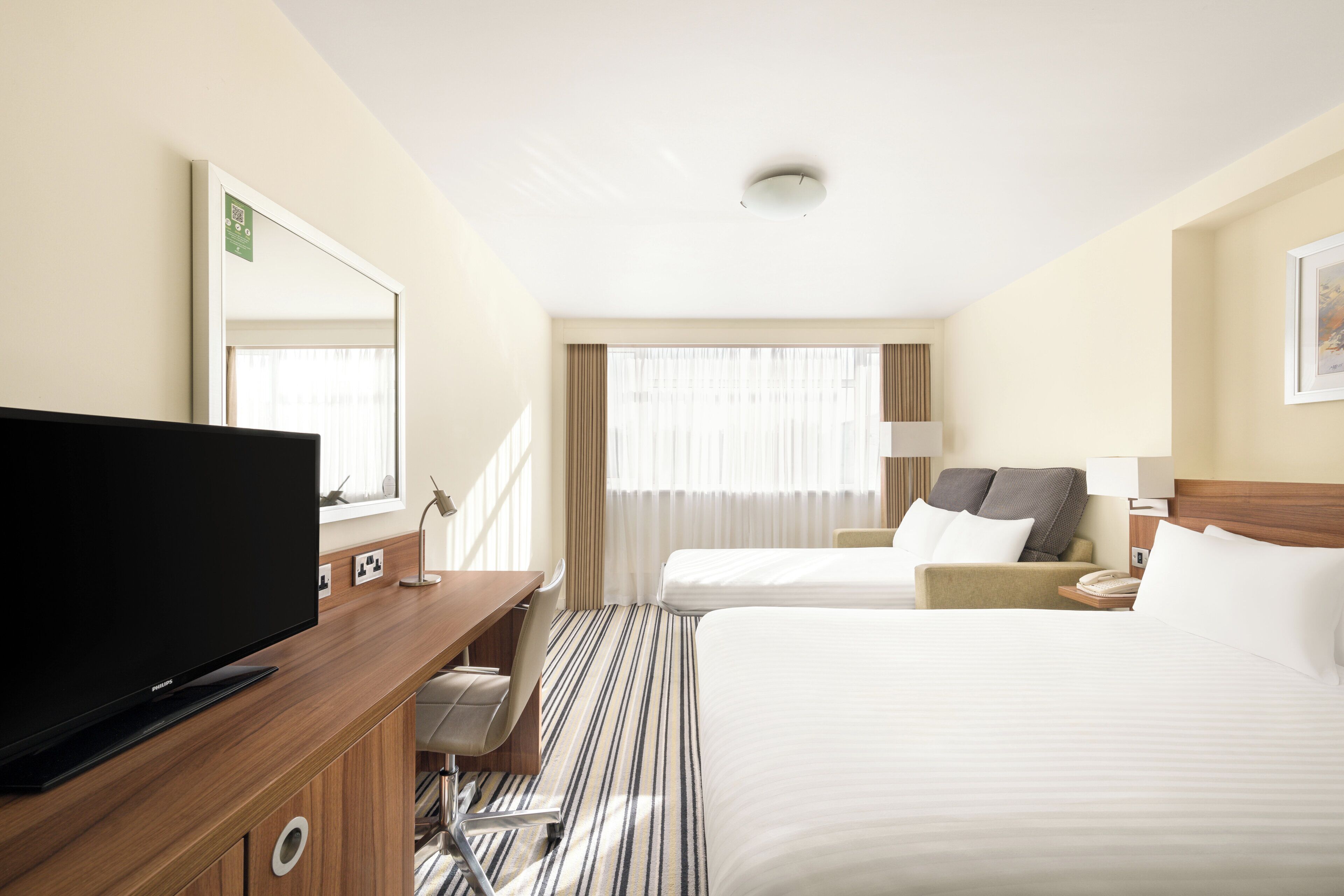 A hotel room with a bed, a desk with a flat-screen TV, a chair, a bedside table with a lamp, and a window with curtains.