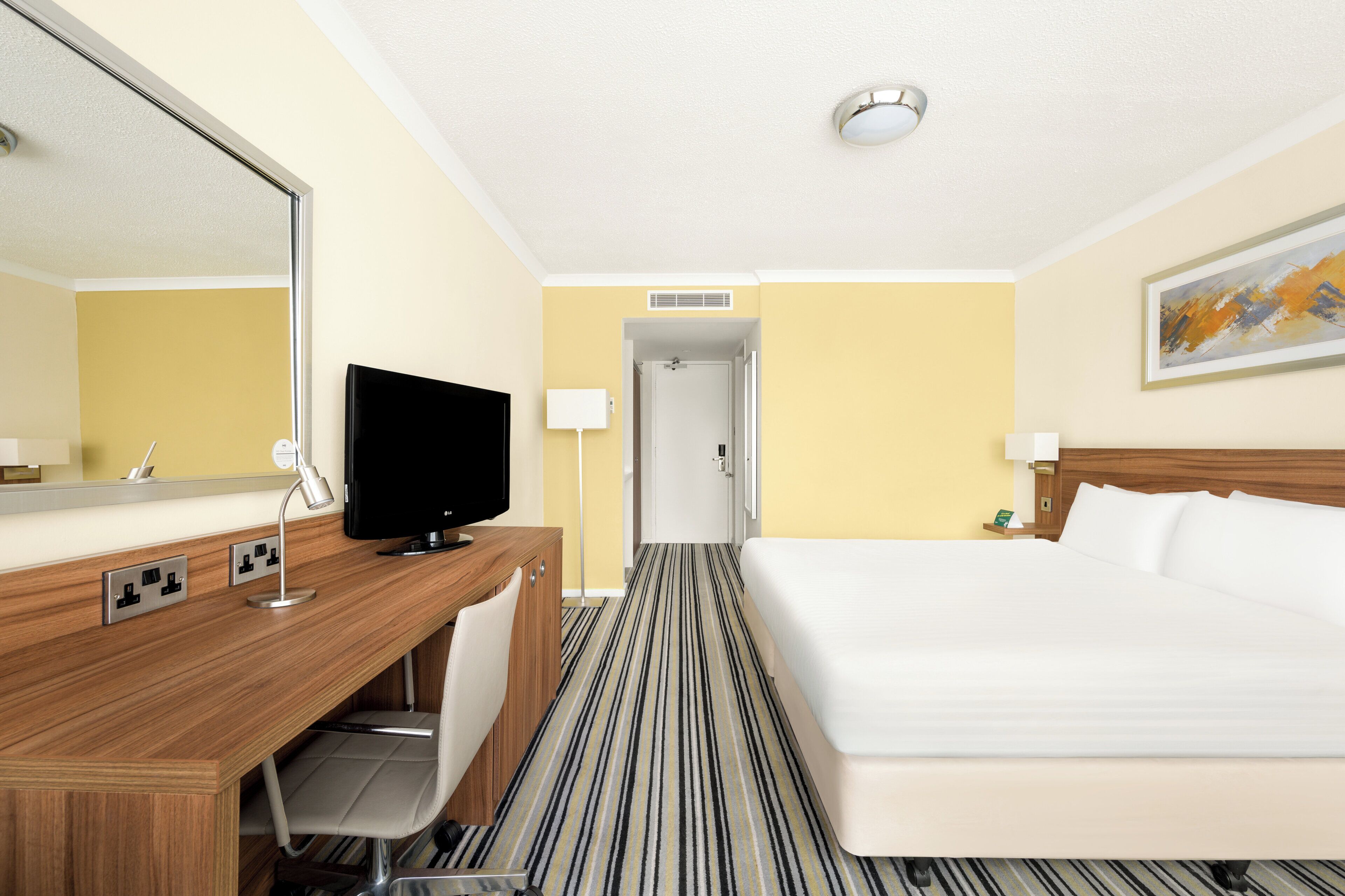 A hotel room with a bed, a desk with a TV, a chair, and a large mirror.
