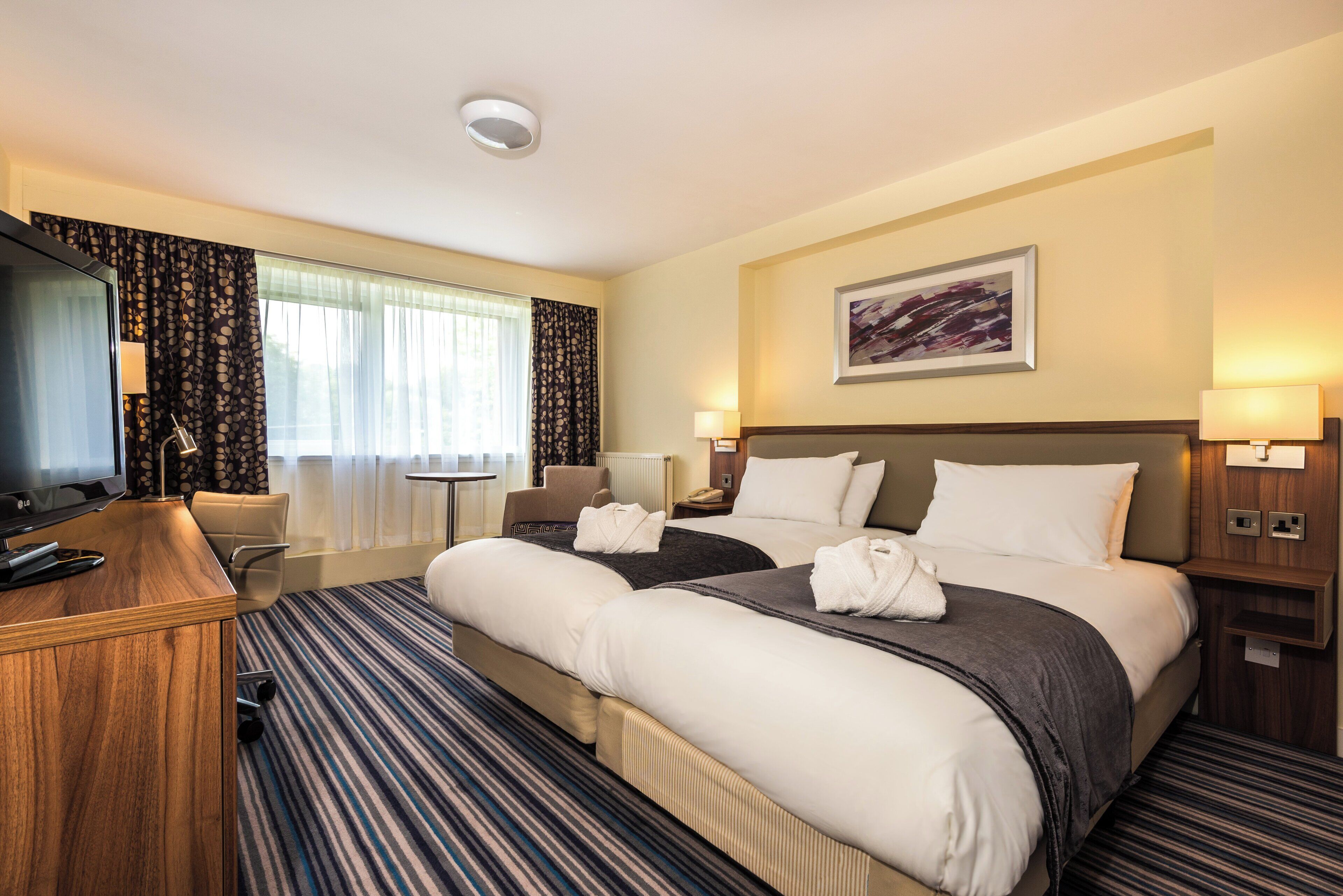 A hotel room with a large bed, a desk, a chair, a TV, and a window with curtains.