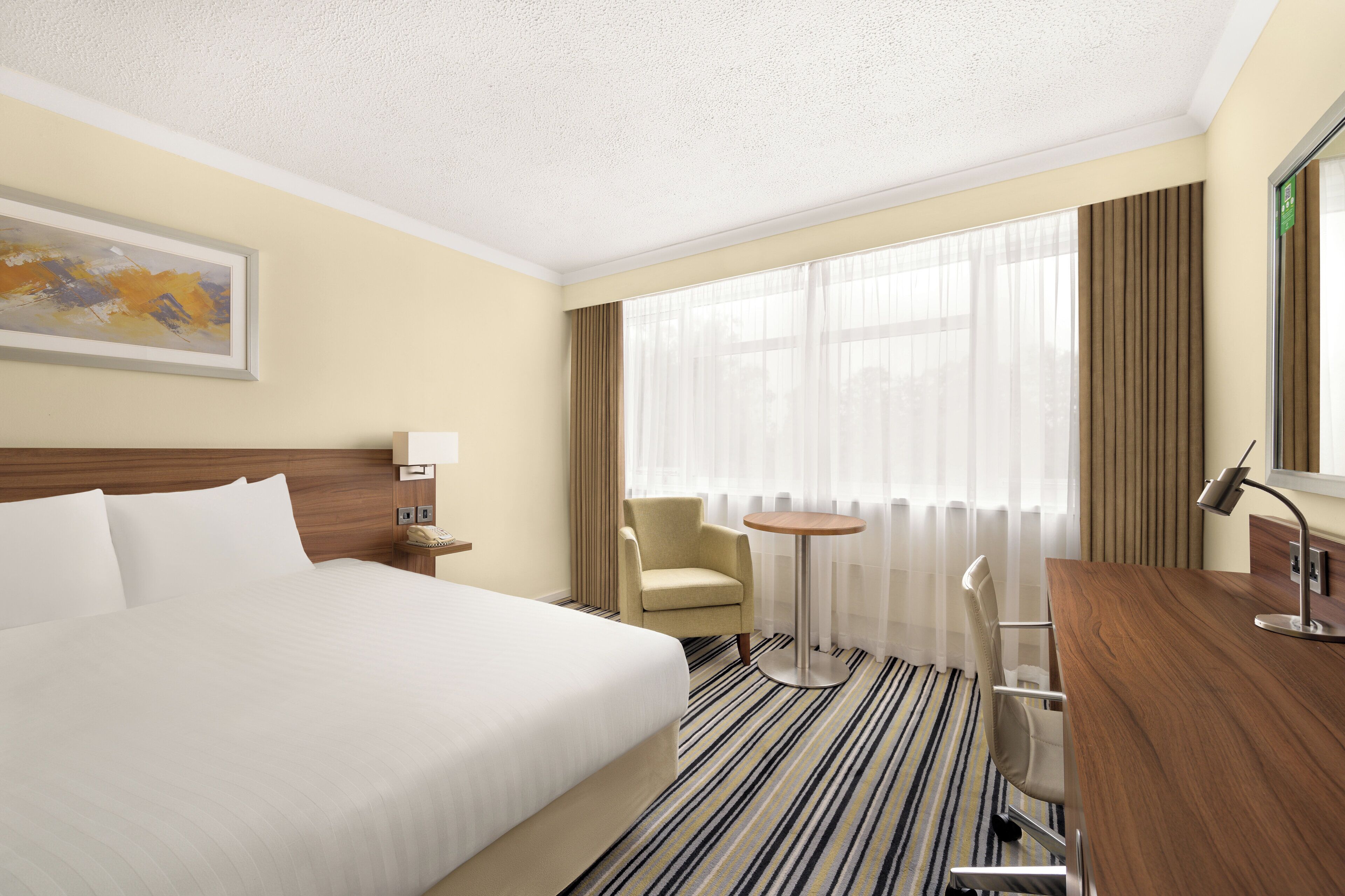 A hotel room with a large bed, a desk, a chair, a small table, and a window with curtains.