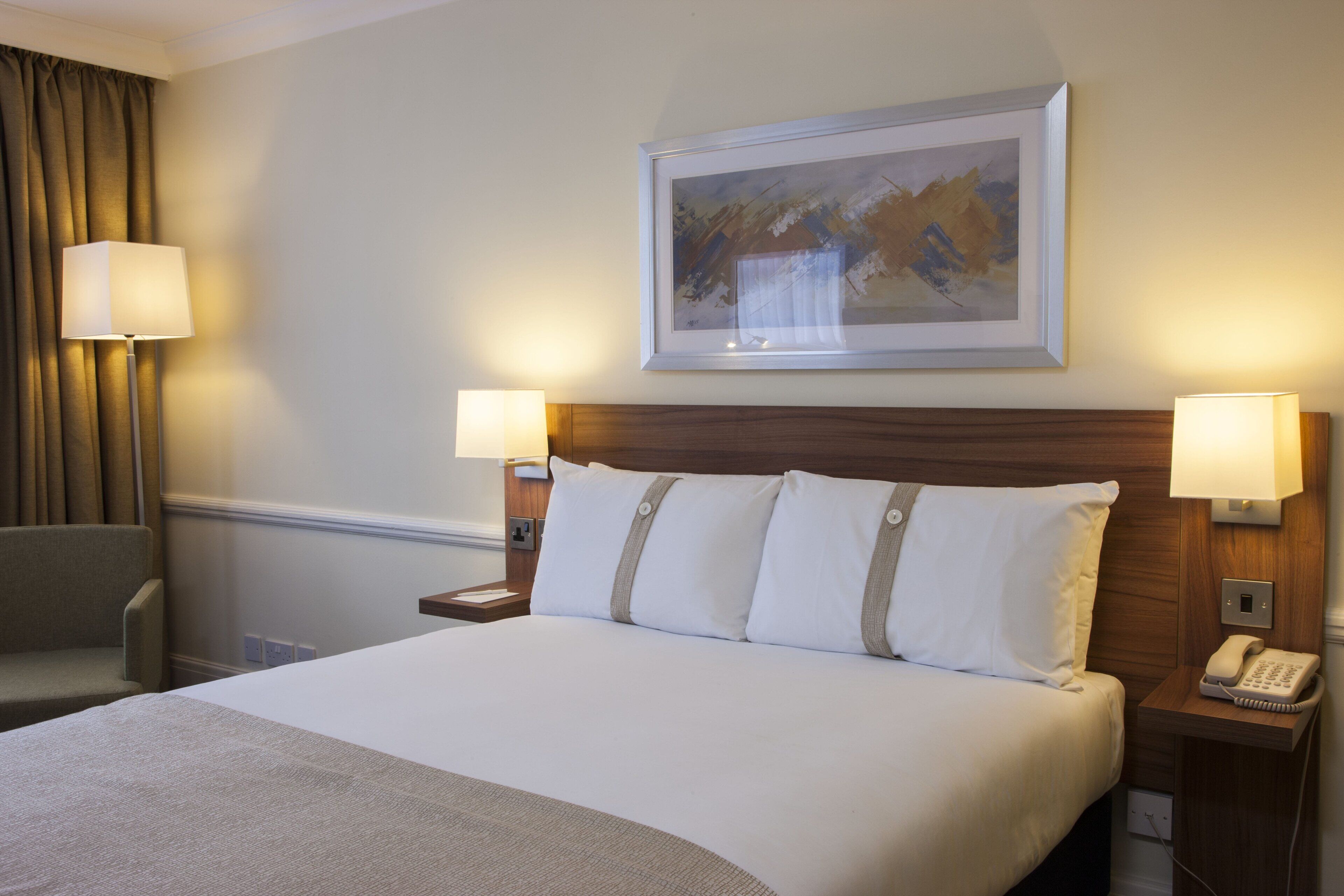 Habitación de hotel con cama, dos lámparas de noche, un cuadro en la pared, una silla y un teléfono en la mesita de noche.
