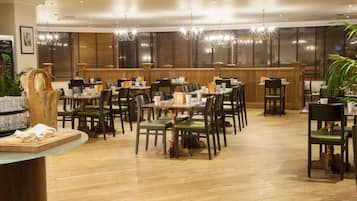Un restaurante con mesas y sillas de madera, una barra con pan y un amplio ventanal con persianas.