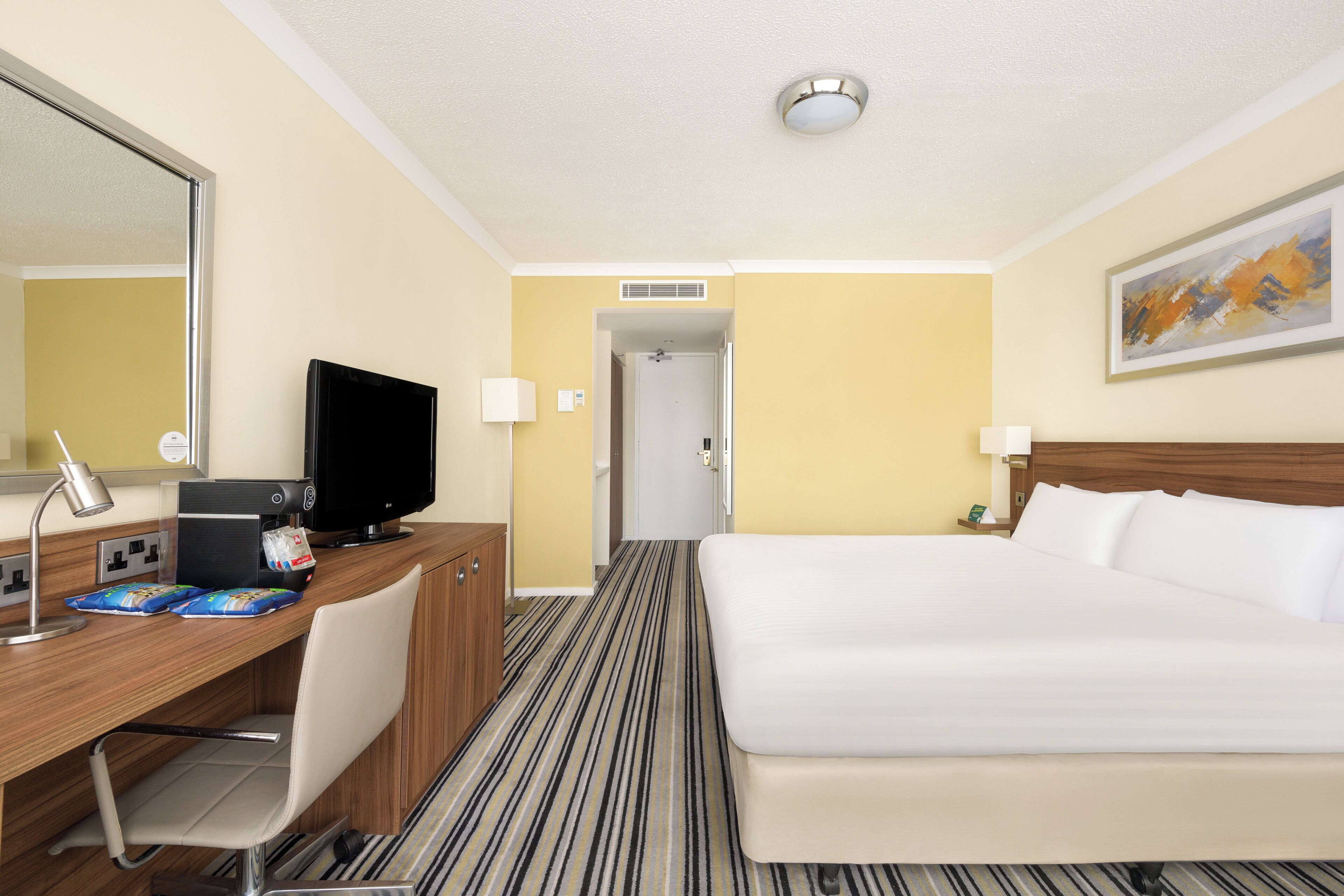 A hotel room with a bed, desk, chair, television, and a painting on the wall.