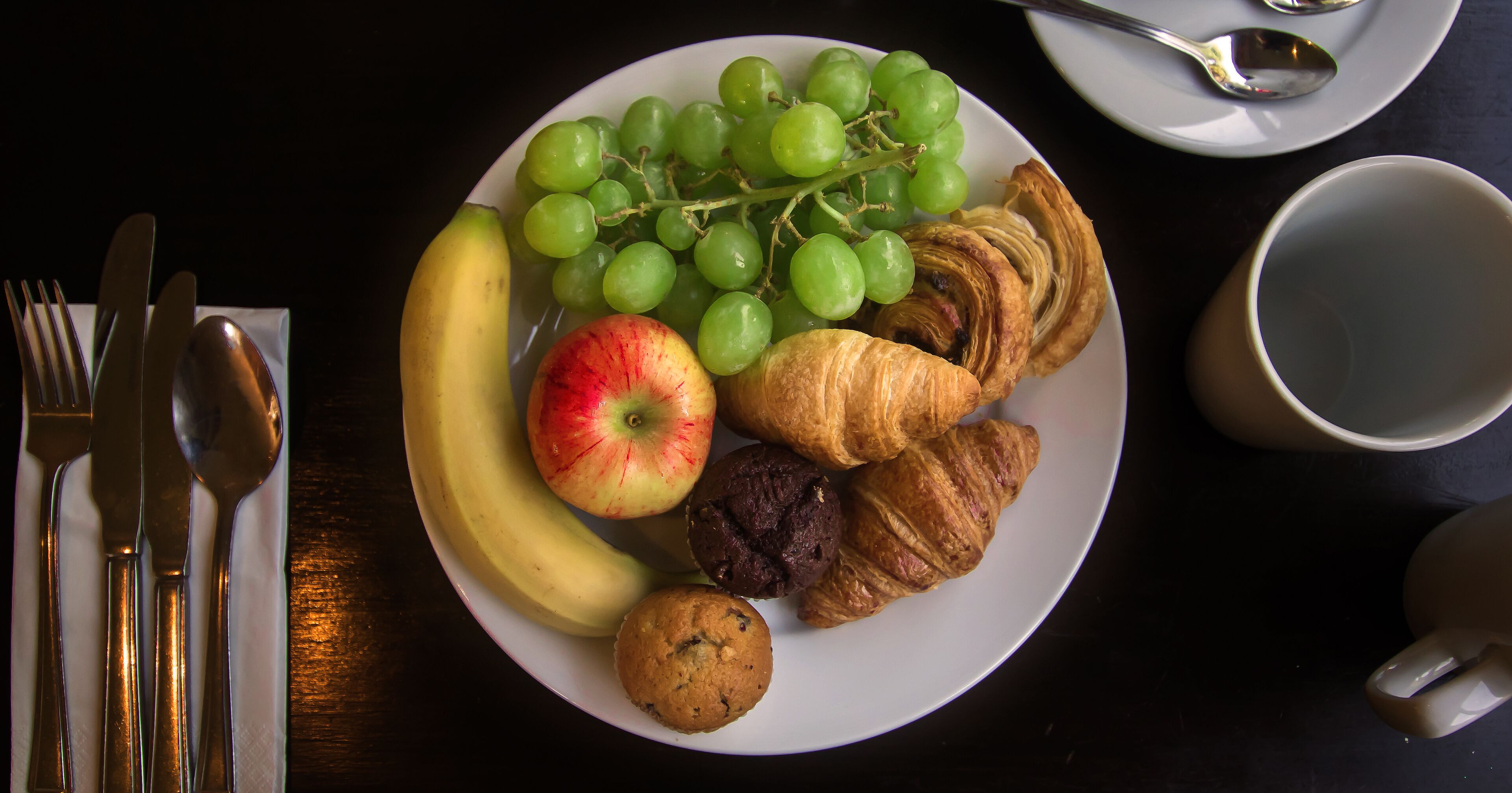 Un plato con plátano, manzana, uvas, croissants y un muffin.