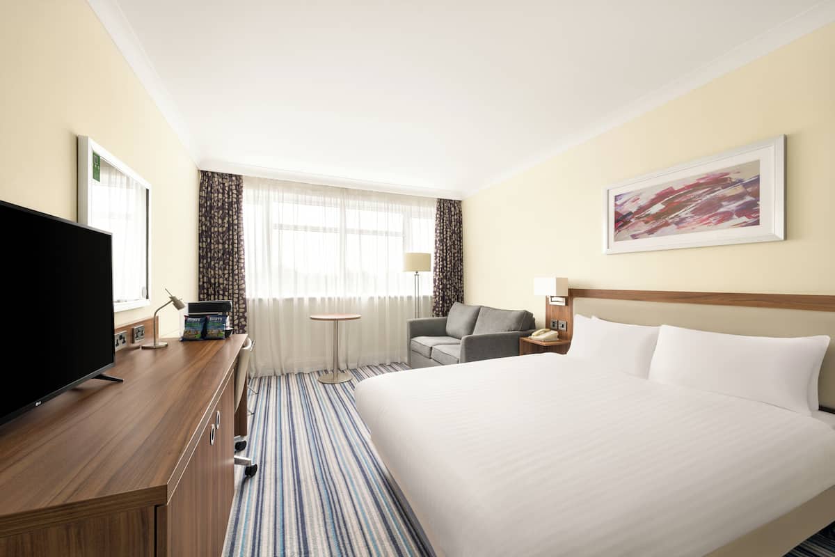 Ein Hotelzimmer mit einem großen Bett, einem Schreibtisch mit Fernseher, einem Sessel, einer Lampe und einem Bild an der Wand.