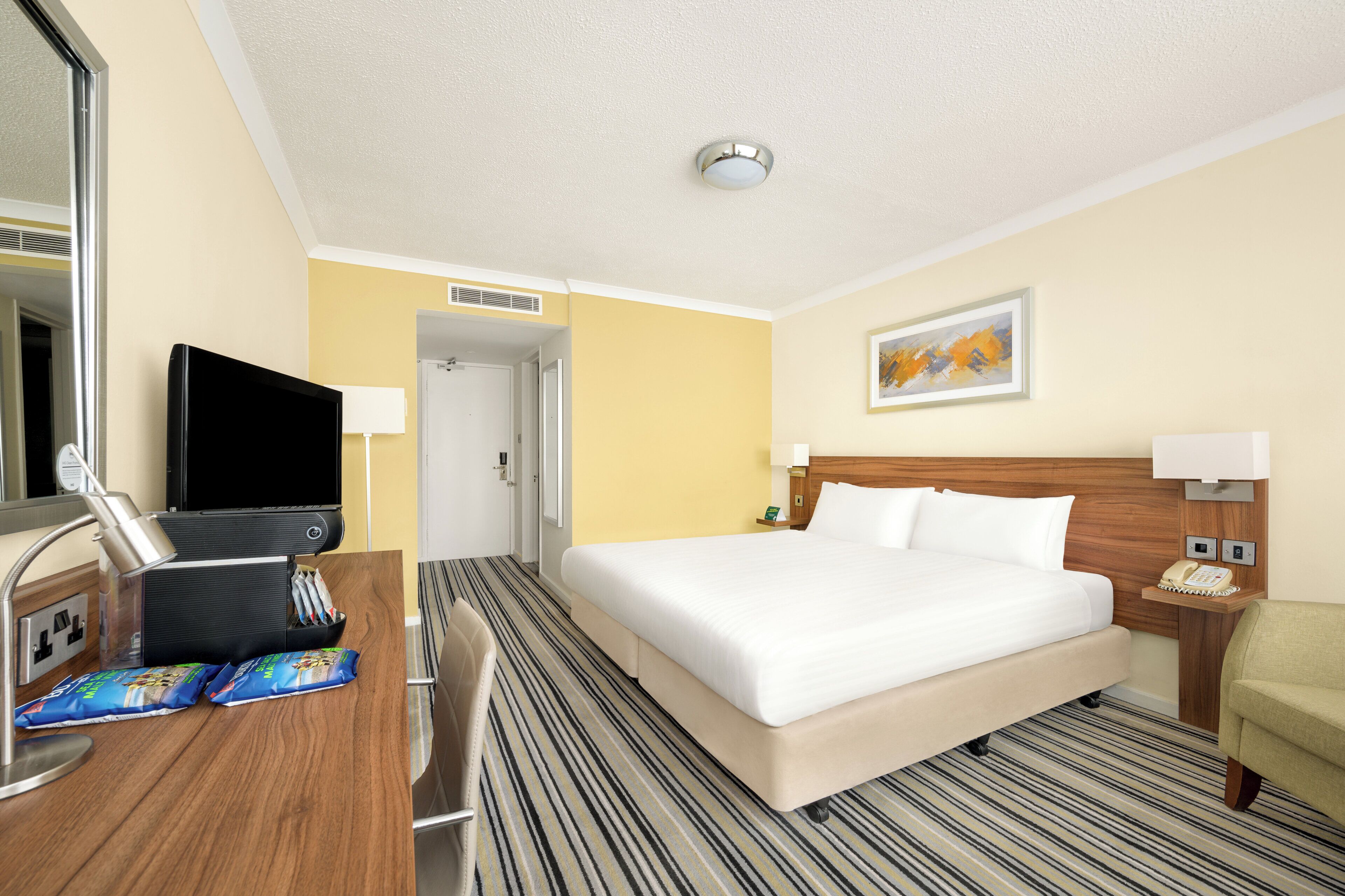 Ein Hotelzimmer mit einem großen Bett, einem Schreibtisch mit Computer und Telefon, einem Sessel und einem Fernseher.