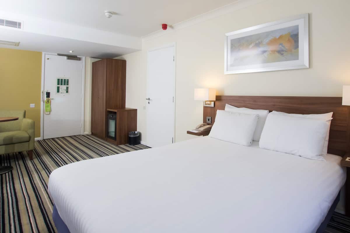 Ein Hotelzimmer mit einem großen Bett, einem Holzschrank, einem Schreibtisch mit Lampe sowie einem Bild an der Wand.