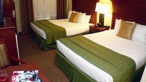 Ein Hotelzimmer mit zwei Betten, jeweils mit grüner Bettdecke und weißen Laken. Zwischen den Betten befindet sich ein Nachttisch mit einer Lampe. Im Vordergrund ist ein Holztisch mit Stuhl und Zeitschrift zu sehen.