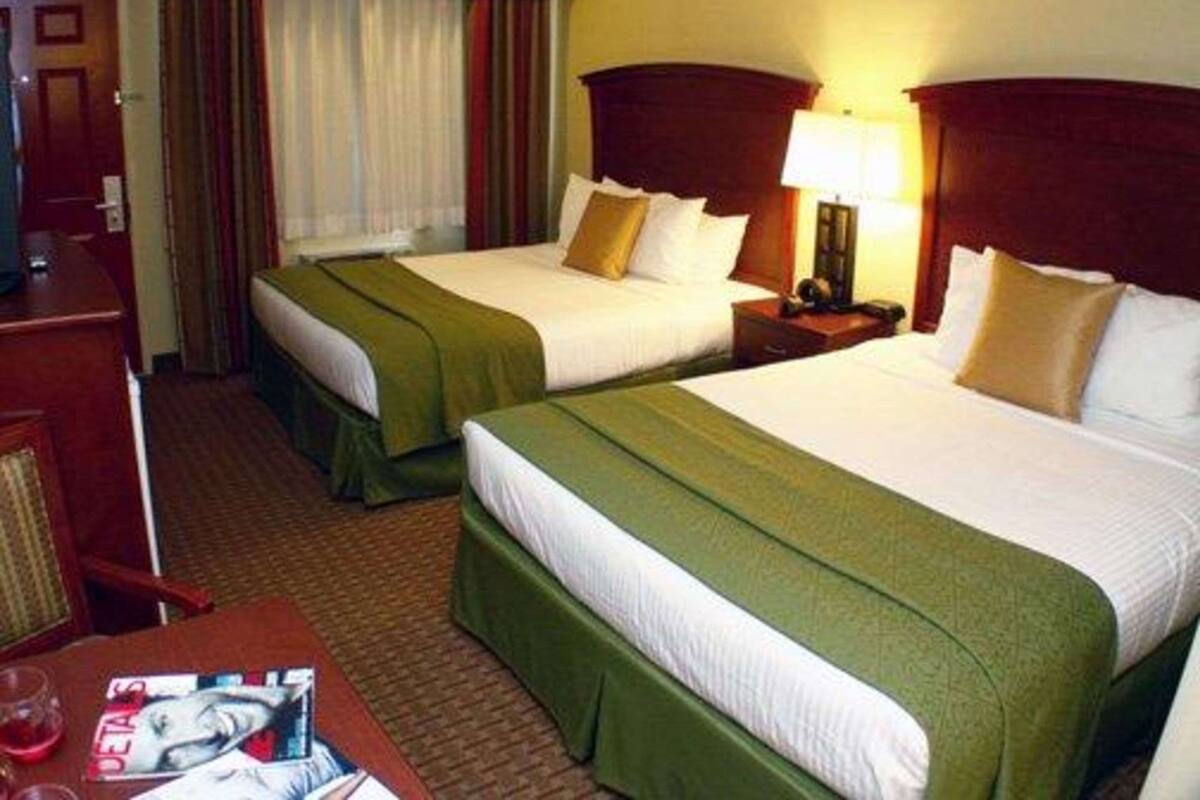A hotel room with two beds, each with a green bedspread and white linens. There is a wooden nightstand with a lamp between the beds. A wooden desk with a chair and a magazine is visible in the foreground.