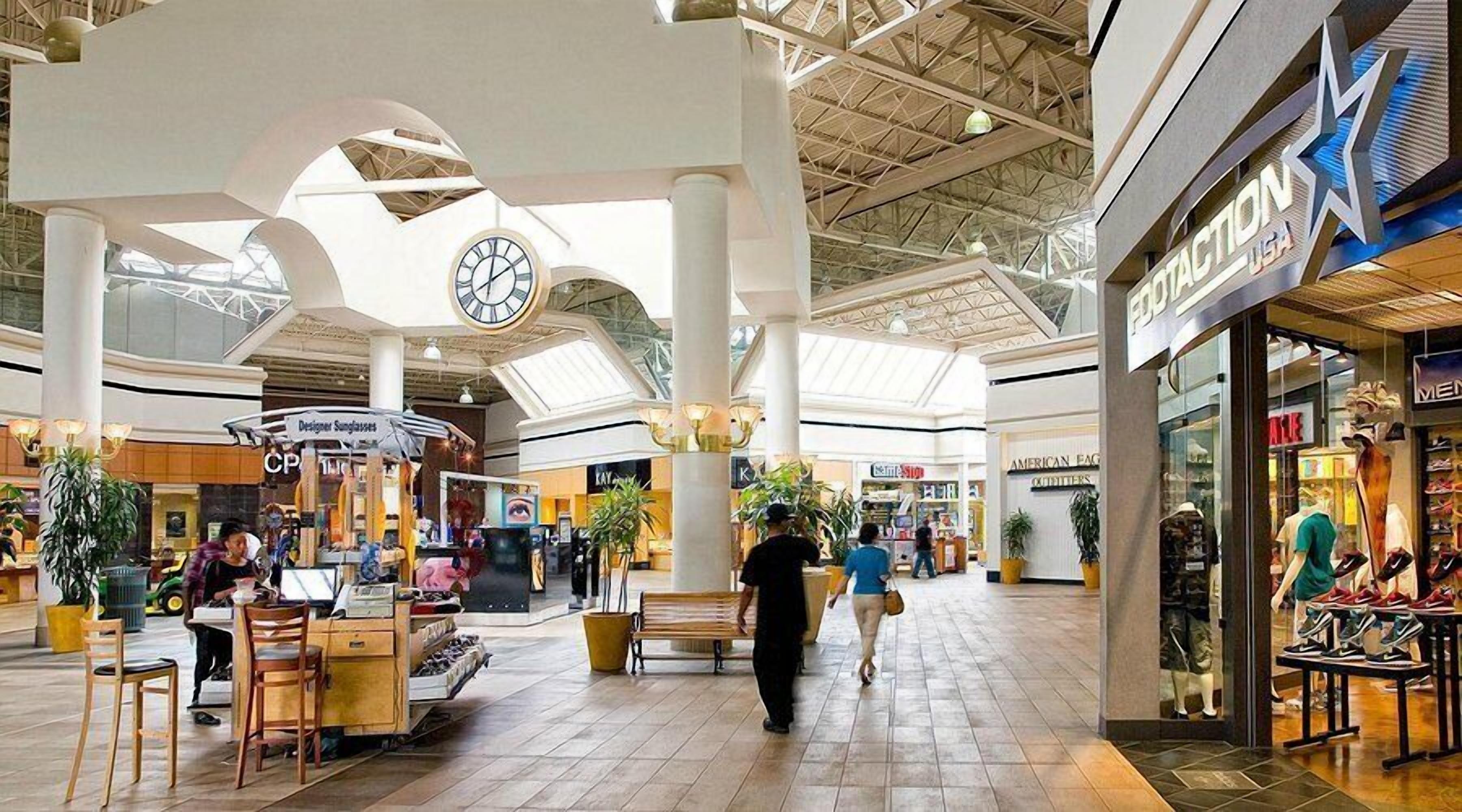 A spacious indoor shopping mall with high ceilings, a central clock, and various retail stores including a shoe store.