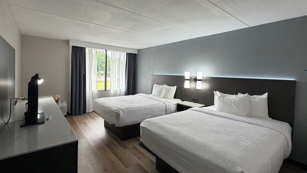 Quarto de hotel com duas camas, uma escrivaninha com abajur e uma janela com cortinas.