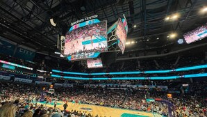 Jogo de basquete em um estádio coberto grande, com várias telas exibindo a partida e placar.