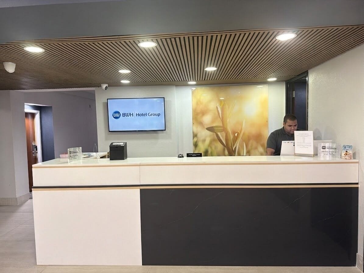 A reception desk with a digital screen displaying the hotel group's name, a man seated behind it, and a modern ceiling design.