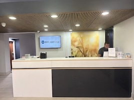 A reception desk with a digital screen displaying the hotel group's name, a man seated behind it, and a modern ceiling design.