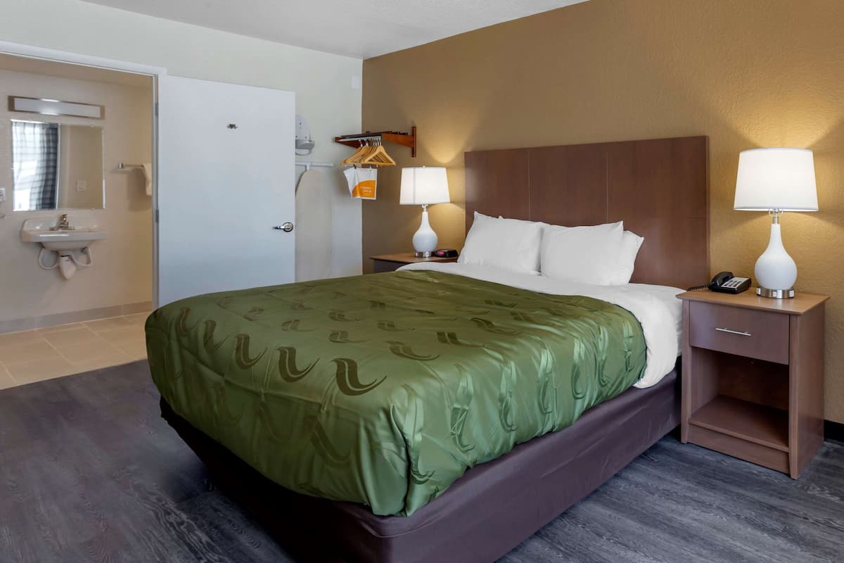 Ein Hotelzimmer mit Bett, zwei Nachttischen mit Lampen, einem kleinen Schrank und einem Badezimmer mit Waschbecken und Spiegel.