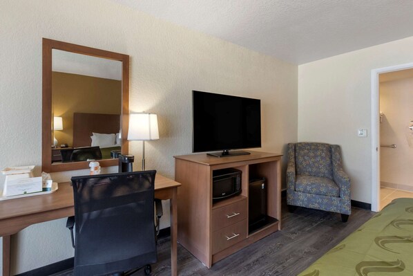 A hotel room with a desk, chair, television, microwave, and a floral armchair.