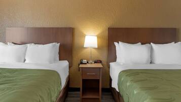 A hotel room with two beds, each with white bedding and green throw pillows. There is a nightstand between the beds with a lamp, a phone, and a clock. The walls are textured and the floor is wooden.