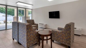 Uma área de espera moderna com cadeiras estampadas, uma mesa redonda e uma TV na parede.
