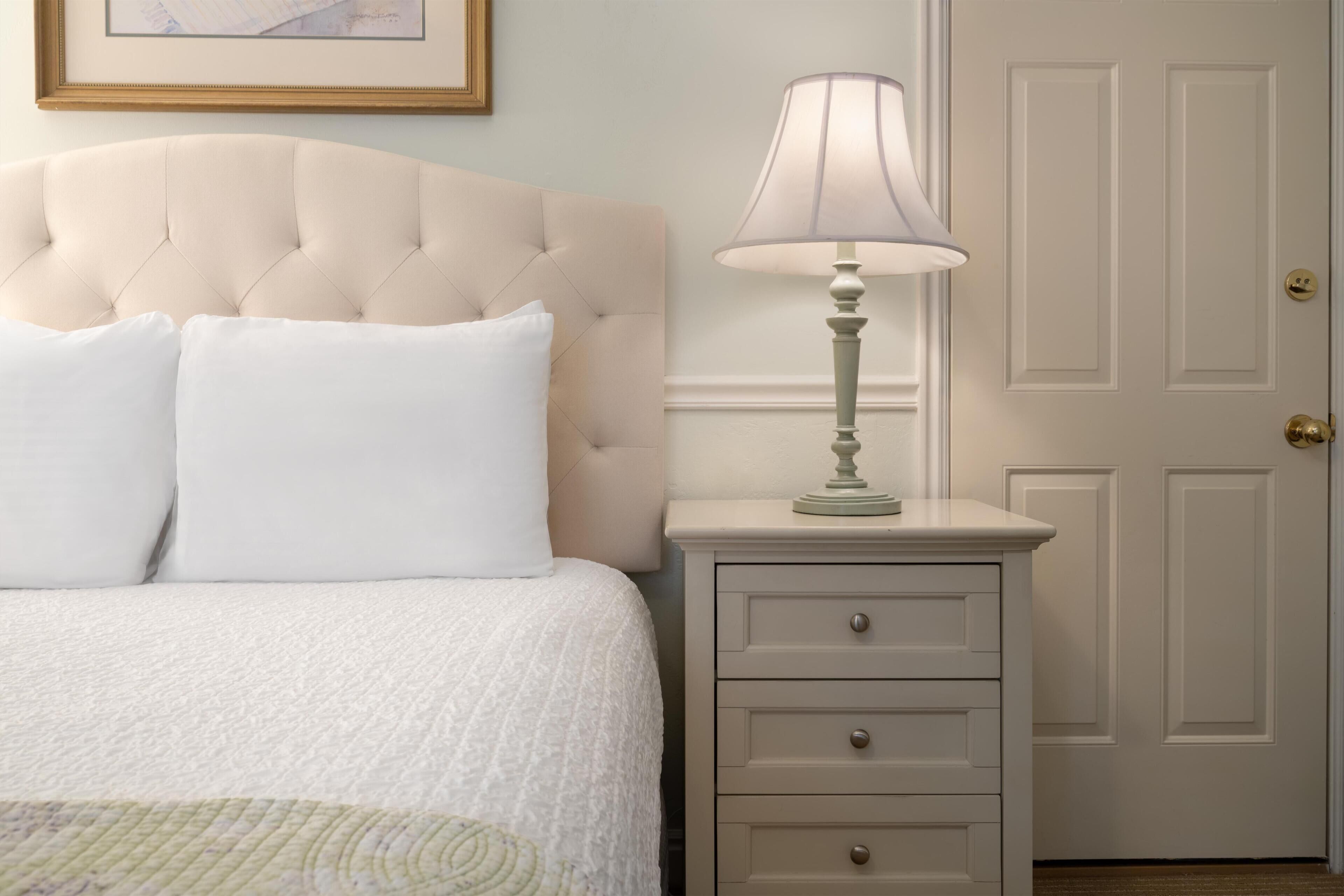 A bedroom with a tufted headboard, a bedside table with a lamp, and a door in the background.