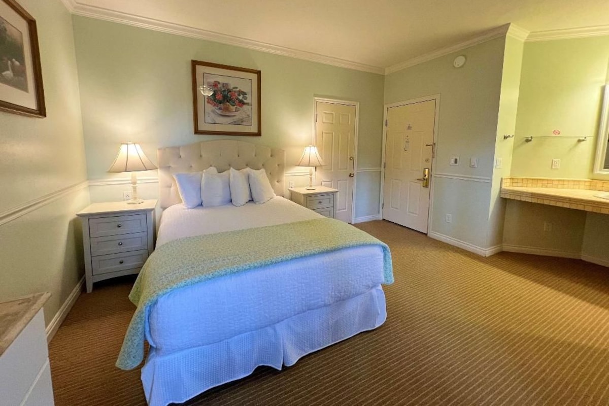 Una habitación de hotel con una cama, dos mesitas de noche, un velador, un ventilador de techo, un espejo adosado a la pared y una puerta.