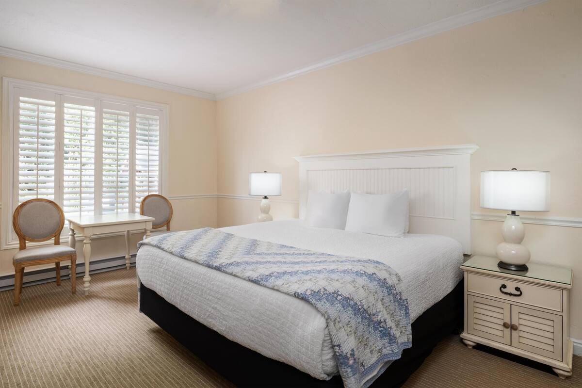 A hotel room with a large bed, two bedside tables with lamps, a desk with a chair, and a window with blinds.