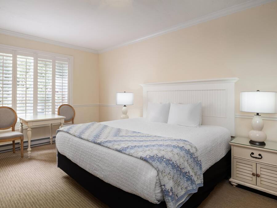 A hotel room with a large bed, two bedside tables with lamps, a desk with a chair, and a window with blinds.