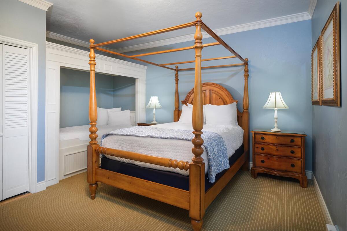 A four-poster bed with a wooden frame and a headboard, a bedside table with a lamp, and a wall-mounted picture frame.