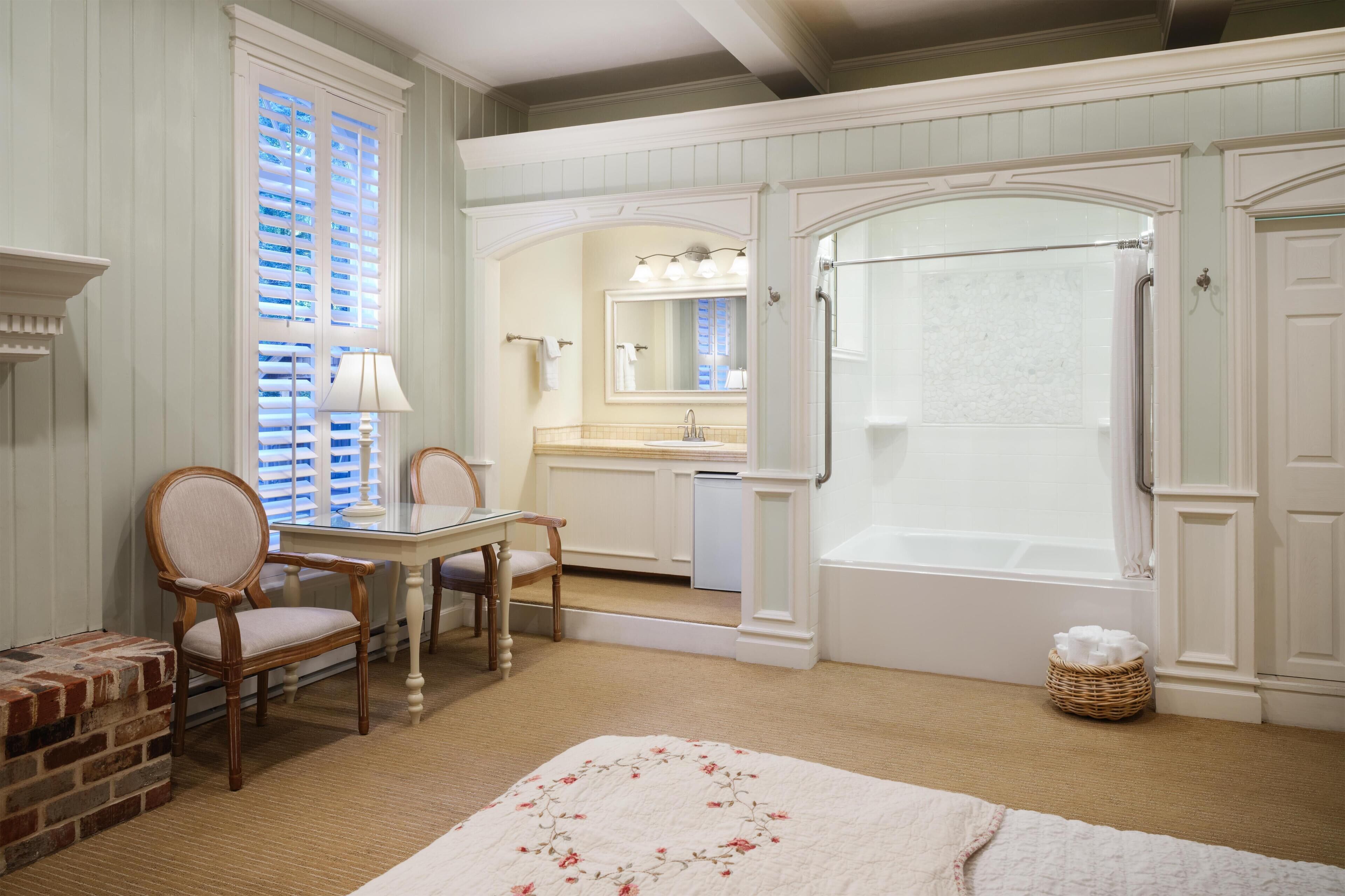 A bathroom with a bathtub, a sink, and a mirror. There is a chair and a table with a lamp.