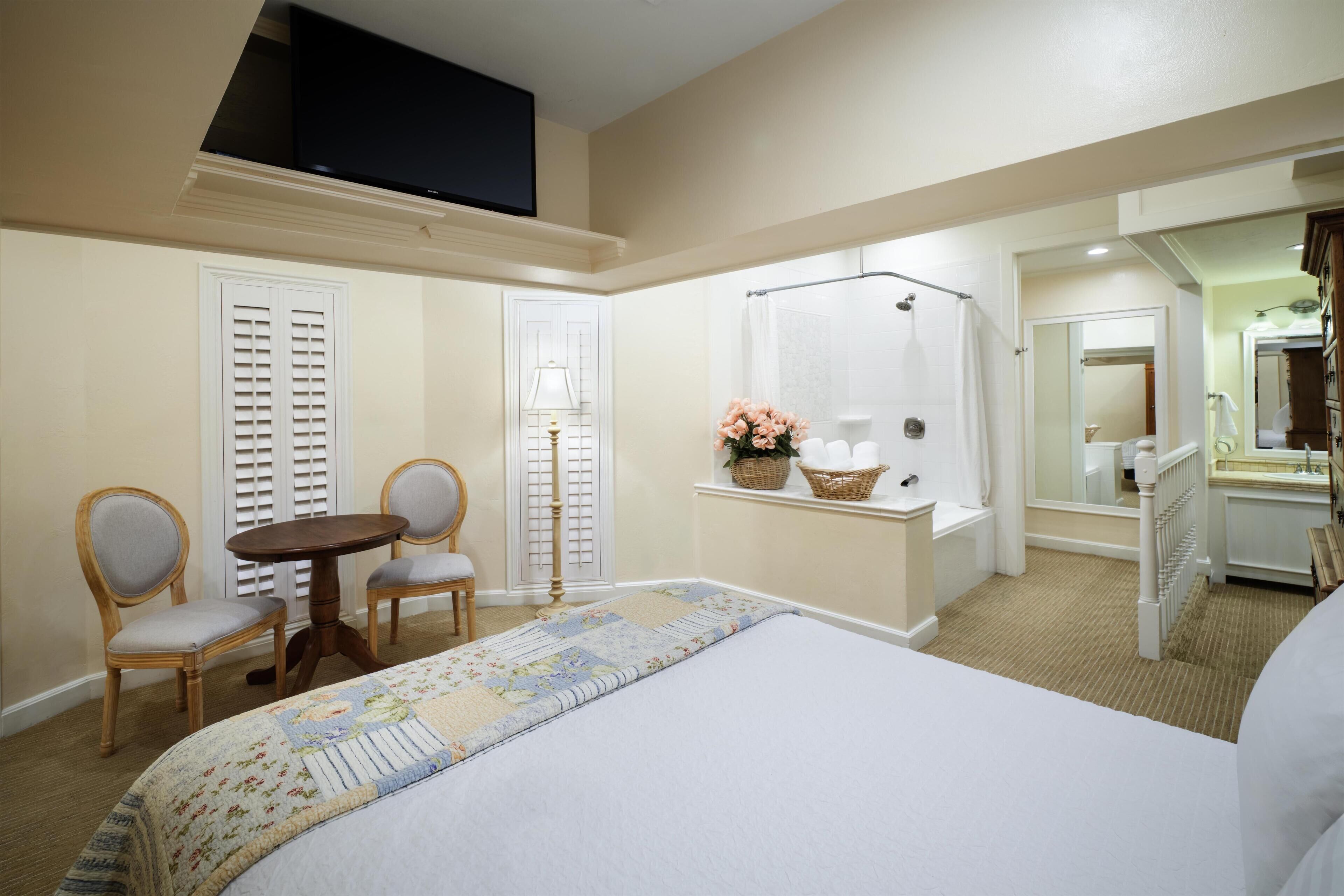 Habitación de hotel con cama, dos sillas, una mesita, televisor, ducha, espejo y ventana con persianas.