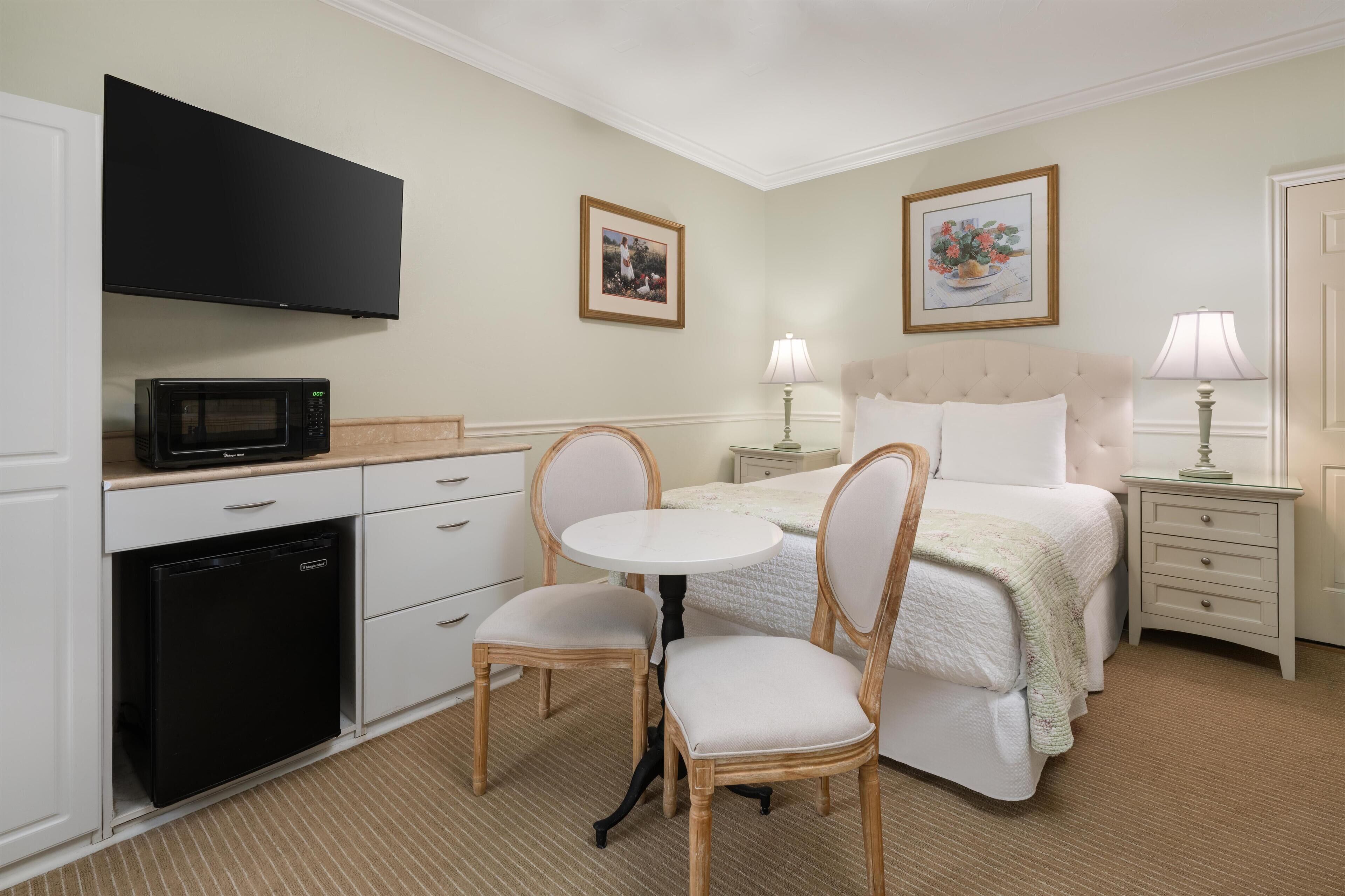 A hotel room with a bed, a TV mounted above a mini-fridge, a dining table with chairs, and two bedside lamps.