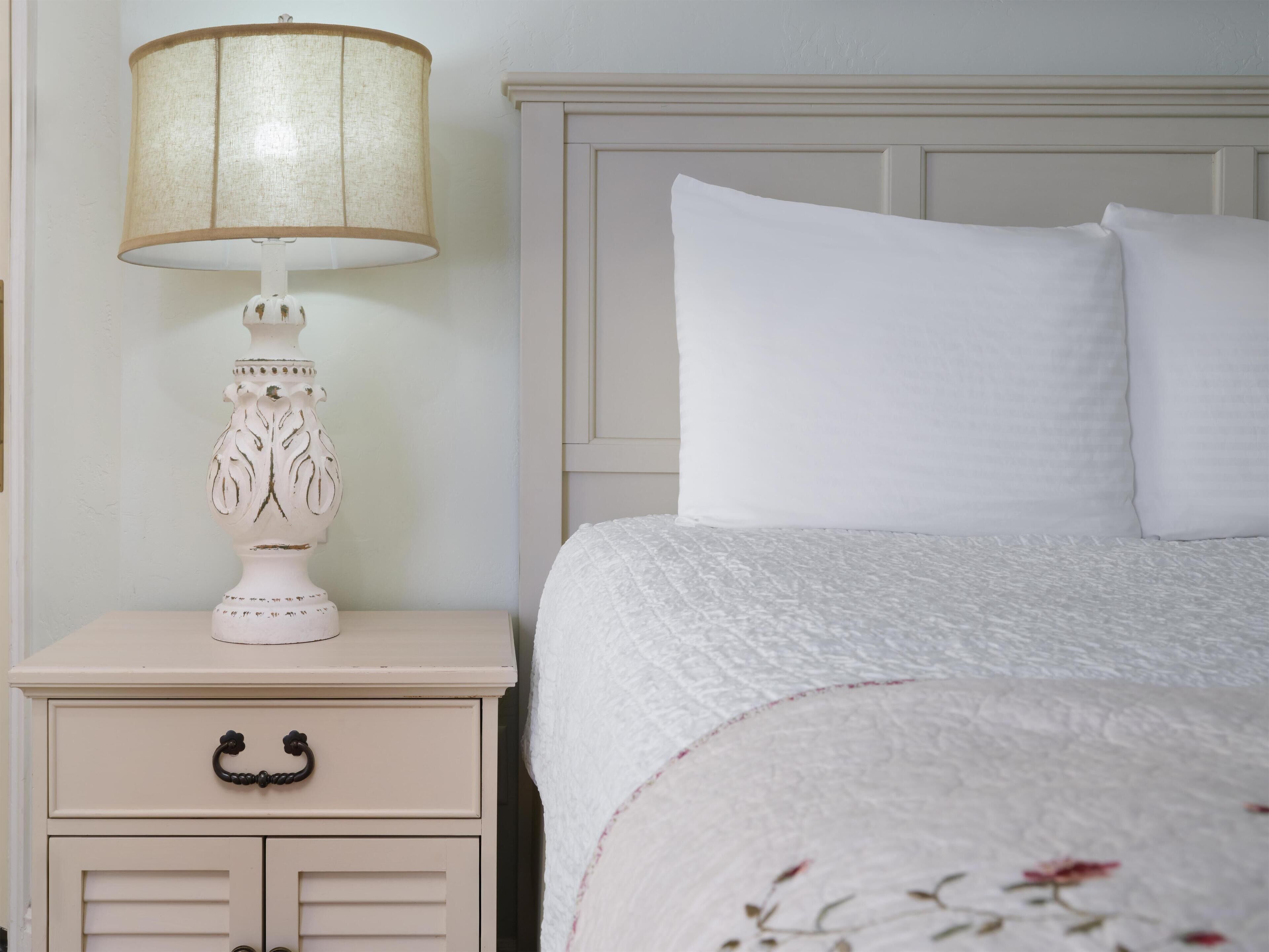 A neatly made bed with white linens and a floral-patterned blanket, a white bedside table with a lamp, and a small drawer with a black handle.