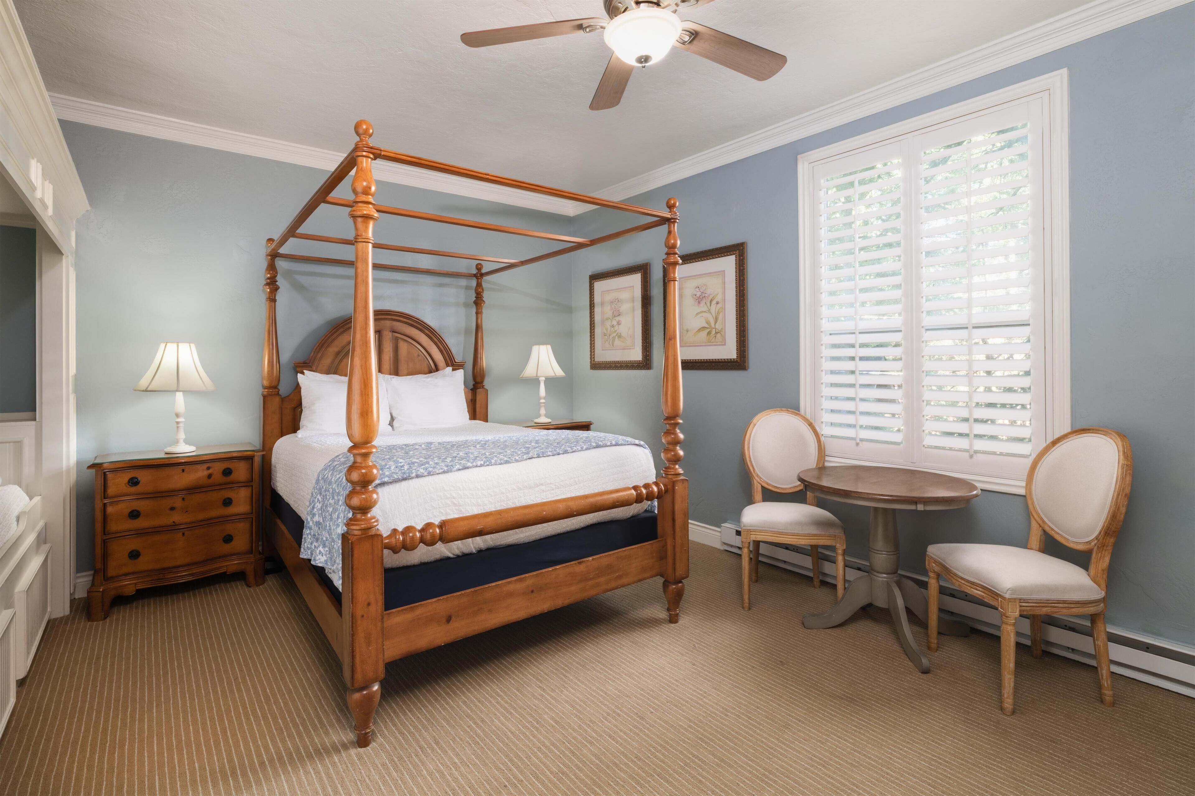 A four-poster bed with a wooden frame, a nightstand with a lamp, a round table with two chairs, and a window with blinds.