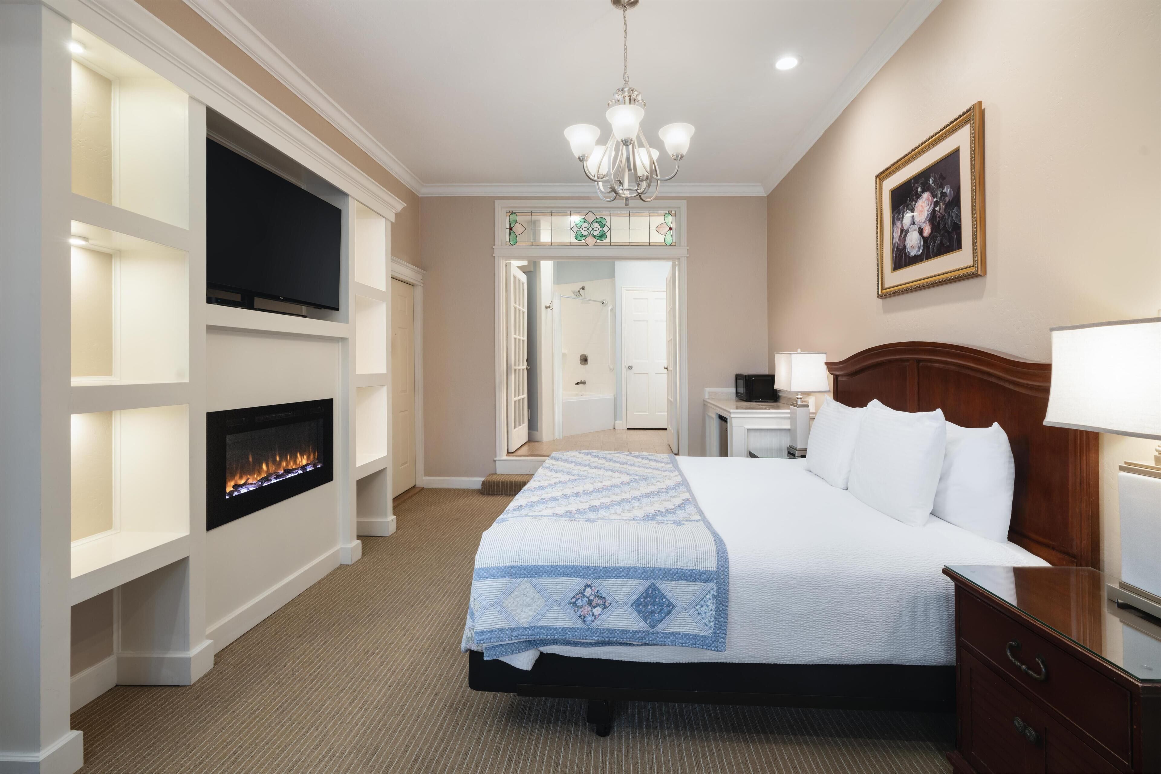 A hotel room with a bed, bedside table, lamp, TV, fireplace, and a door leading to a bathroom.
