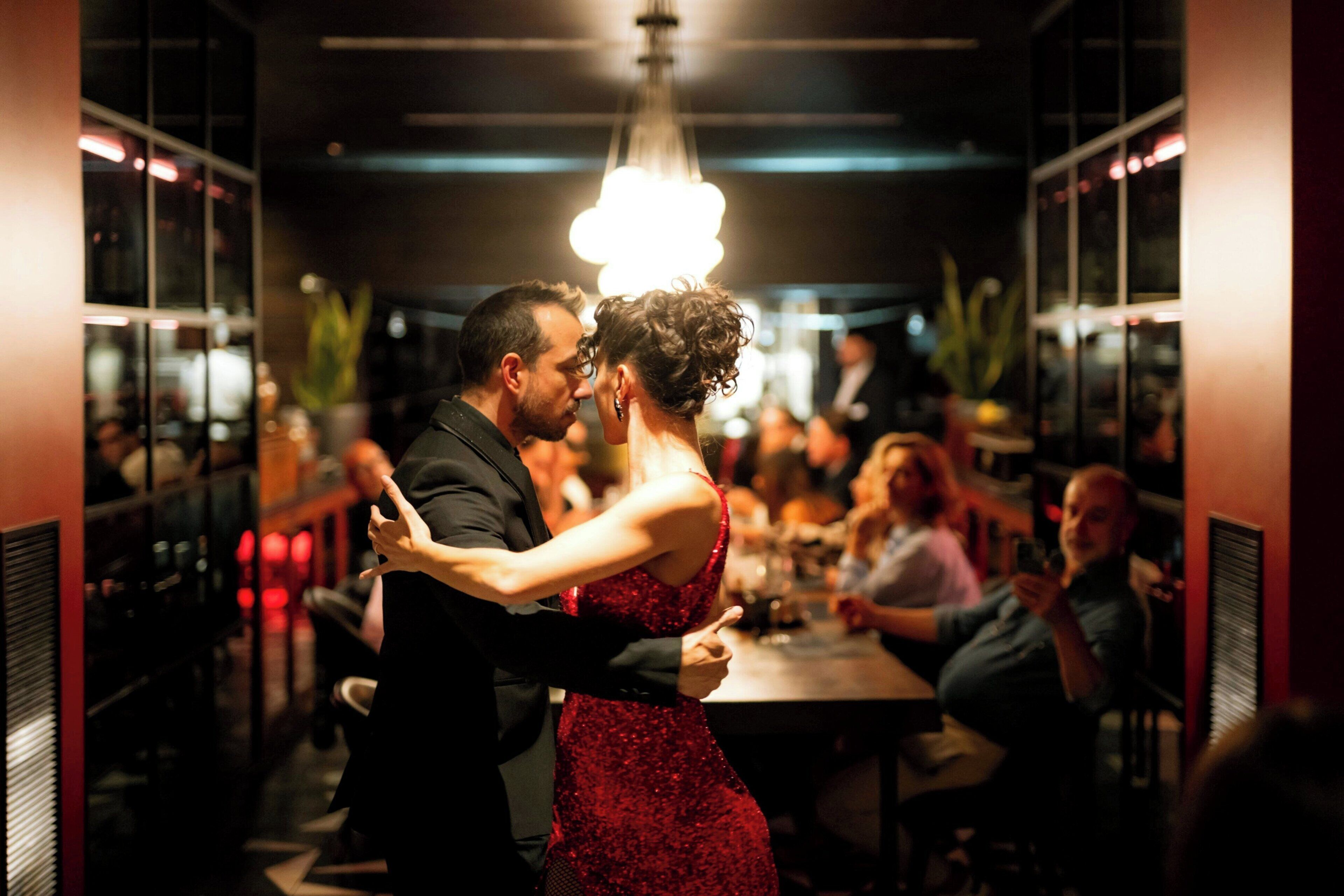 Un couple danse dans une pièce faiblement éclairée, sous un lustre, entouré d’invités assis.