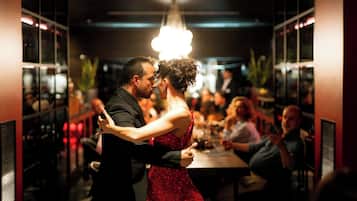 Una pareja baila en una sala tenuemente iluminada con un candelabro, rodeada de invitados sentados.