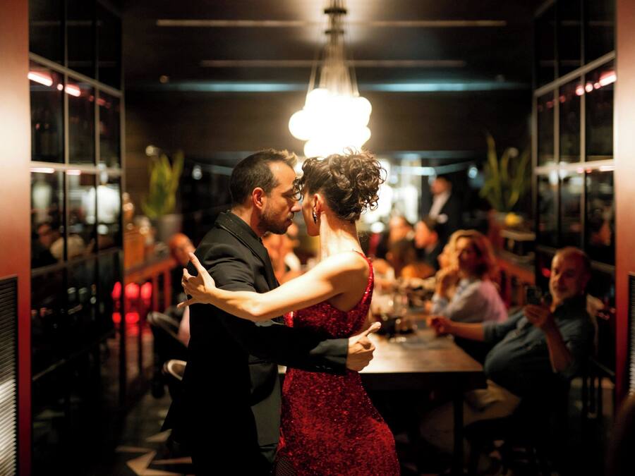 Una pareja baila en una sala tenuemente iluminada con un candelabro, rodeada de invitados sentados.