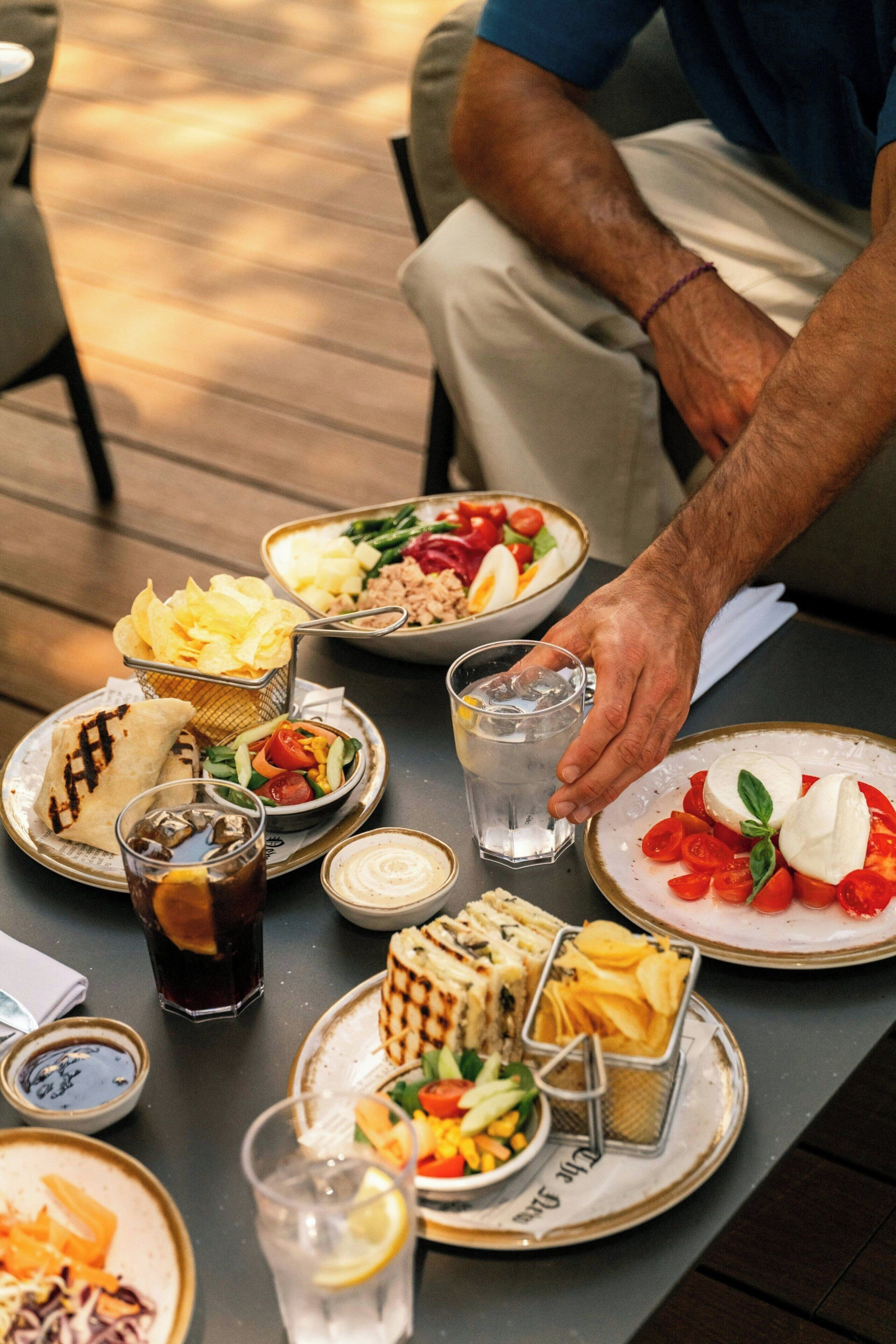 Une table à manger avec différents plats, un verre de boisson fraîche et une personne qui tend la main vers un verre.
