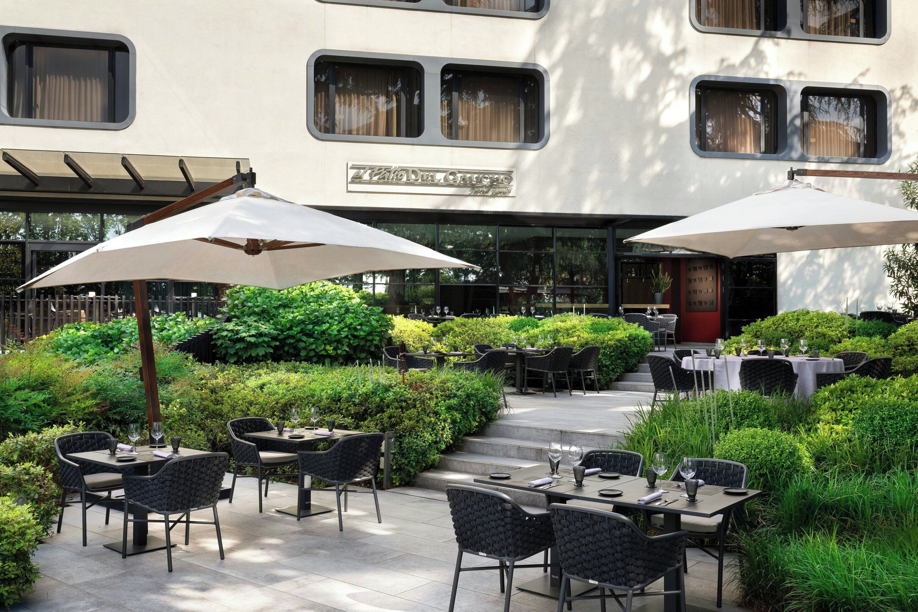 An outdoor dining area with tables and chairs, surrounded by greenery and a building with large windows.