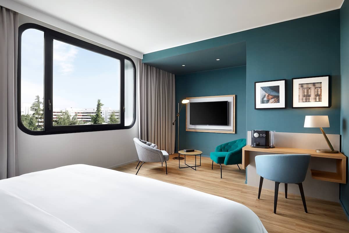 A modern hotel room with a large bed, a desk with a computer, a chair, a small table, and a TV.