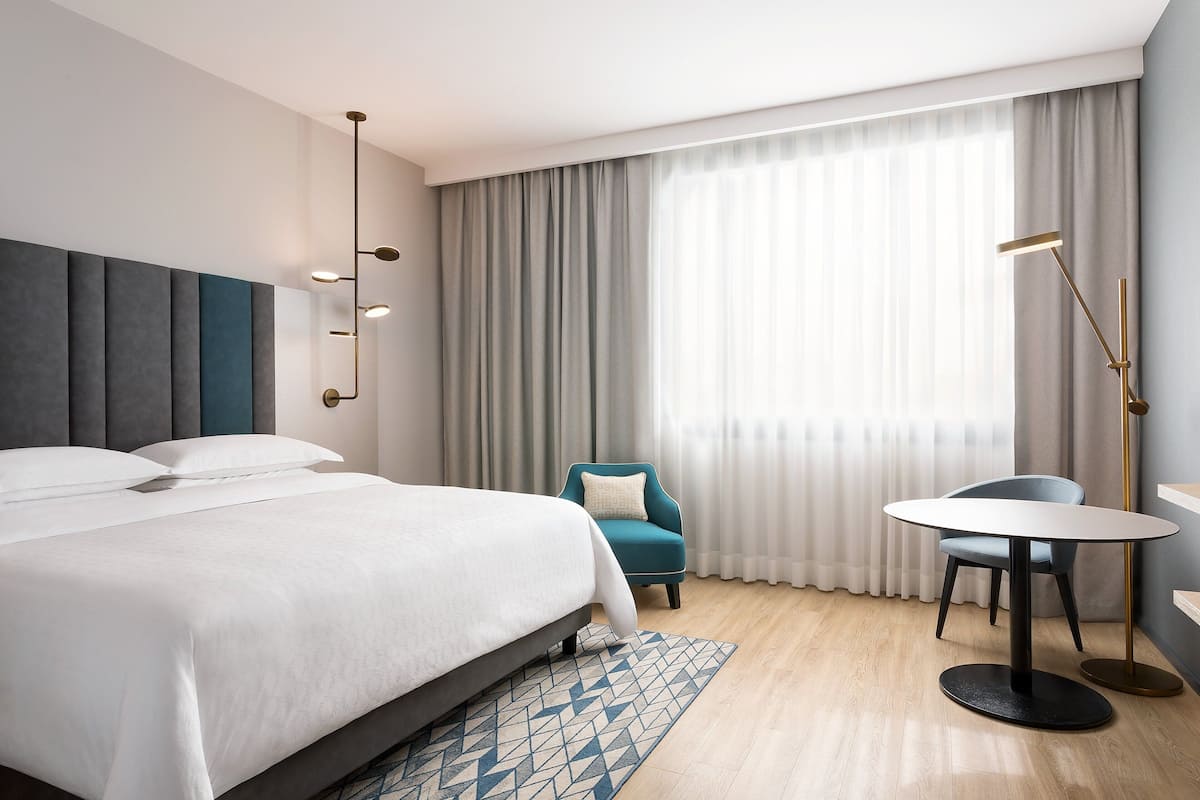 A modern hotel room with a large bed, a small round table, a chair, and a floor lamp.