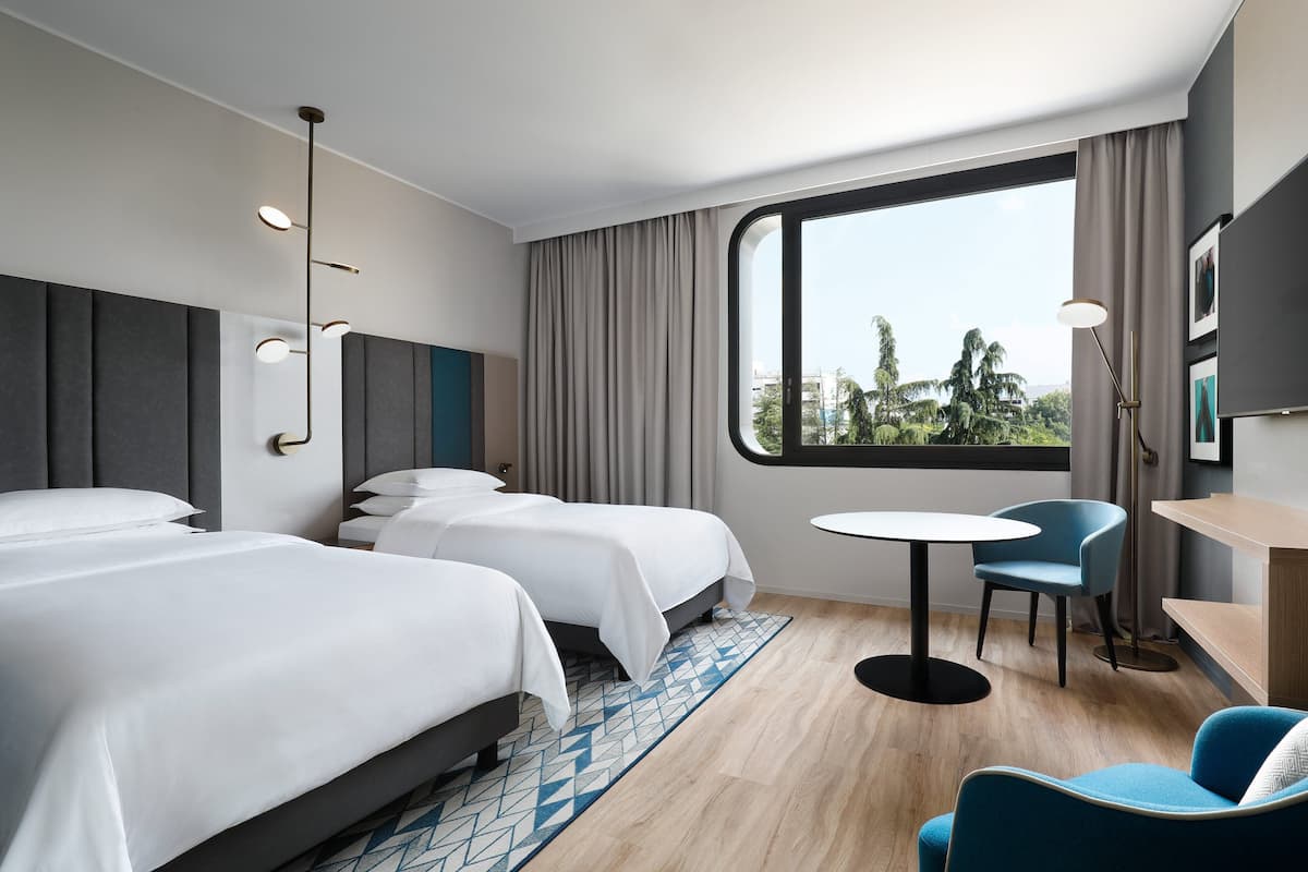 A hotel room with two beds, a round table, a chair, a desk, and a large window with a view of greenery.